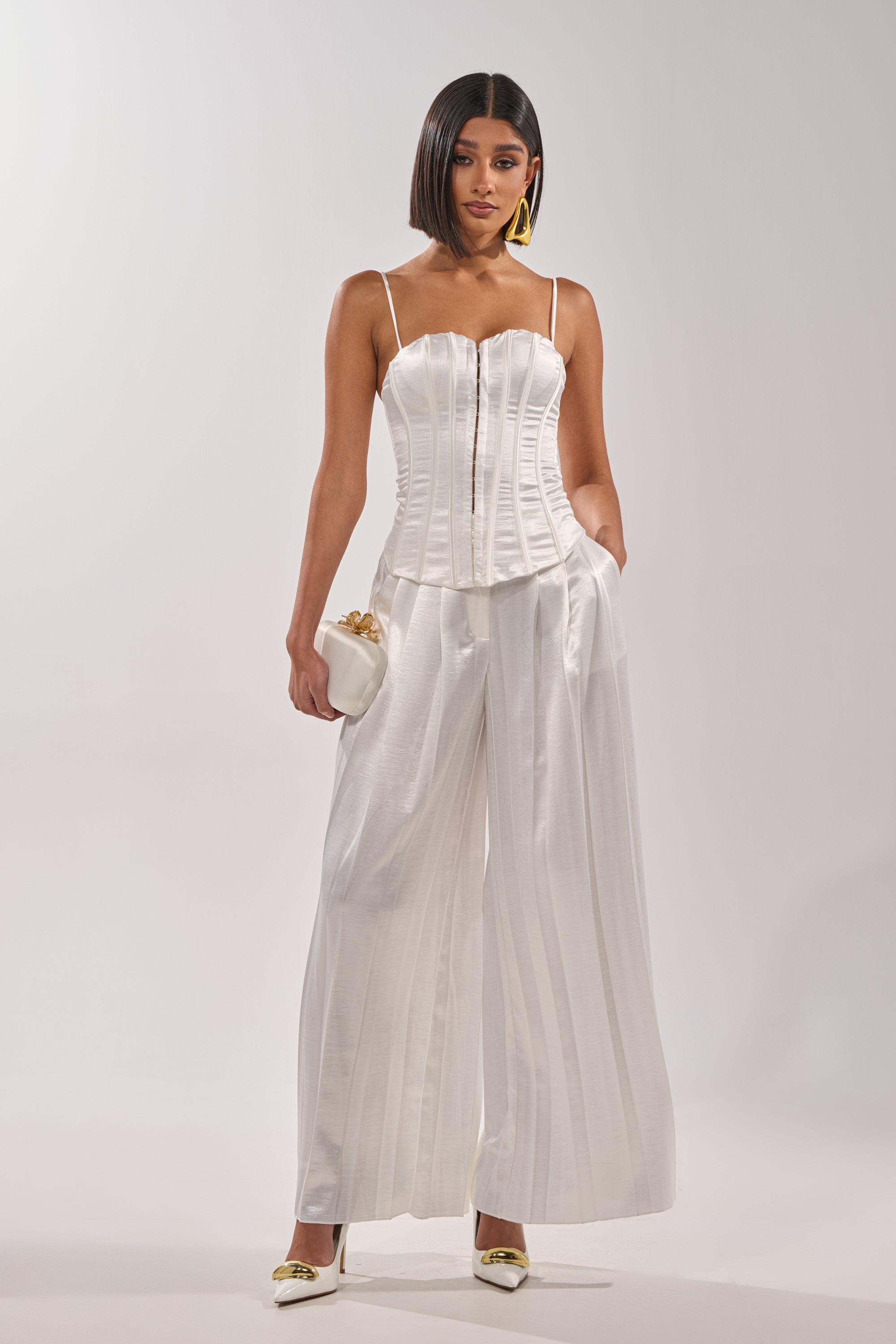 Wearing the IRIS SATIN CORSET with a sweetheart neckline and wide-leg pants, a woman stands with one hand in her pocket, styled with a small white clutch, white heels, statement earrings, and a sleek bob. 4