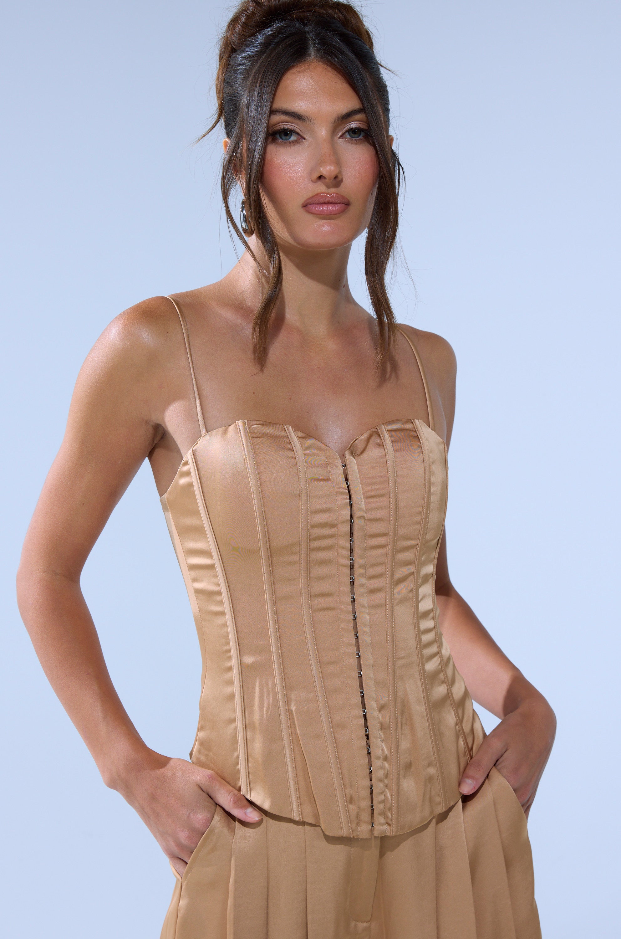 A woman stands with her hands in her pockets against a pale blue background, wearing the HANSEL MOCHA SATIN CORSET with thin straps and matching high-waisted pants.