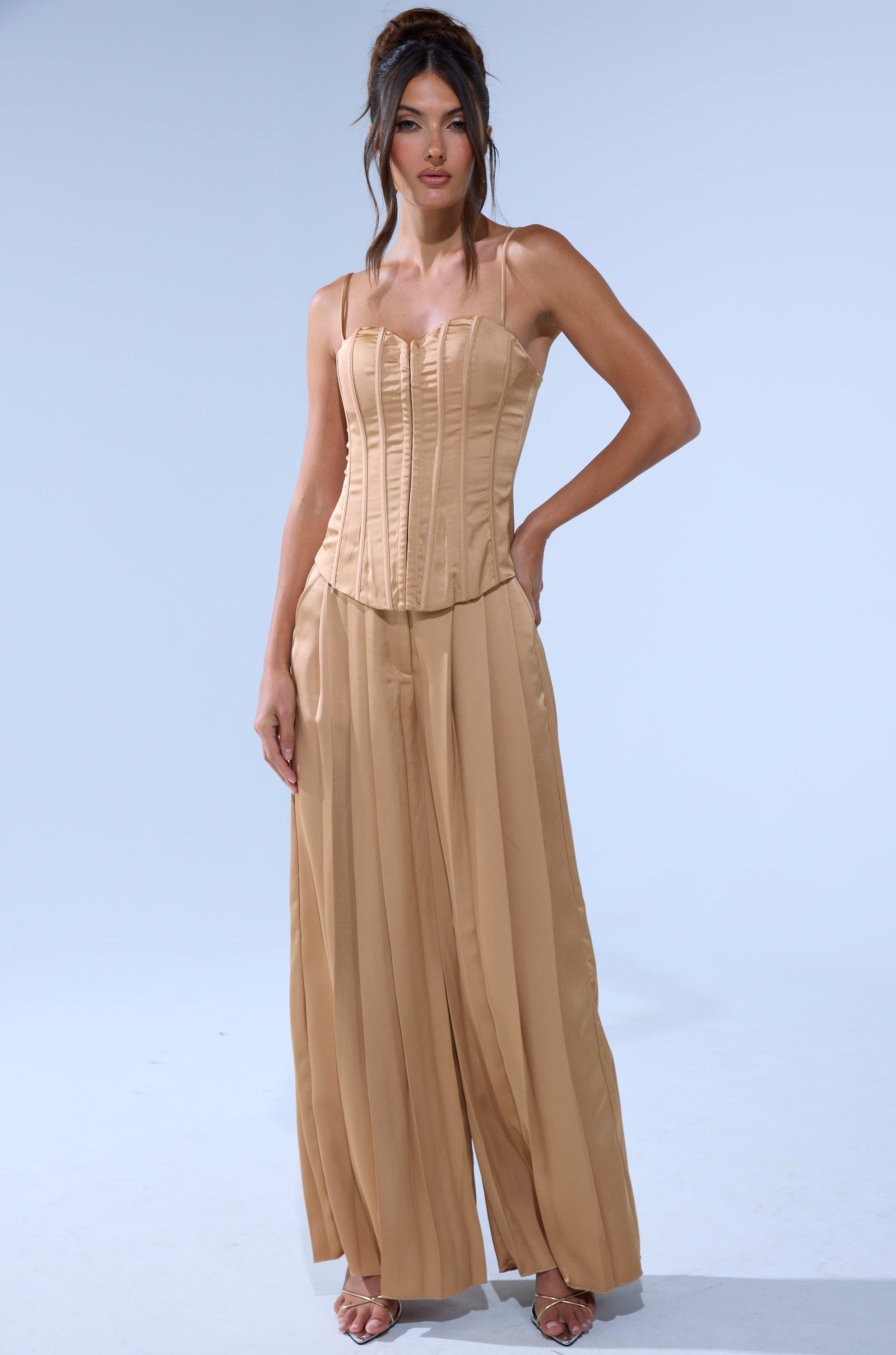 A woman poses against a plain background wearing the HANSEL MOCHA SATIN CORSET with thin straps, paired with matching high-waisted, wide-leg pleated pants. She completes the look with heeled sandals and a high updo featuring loose strands.
