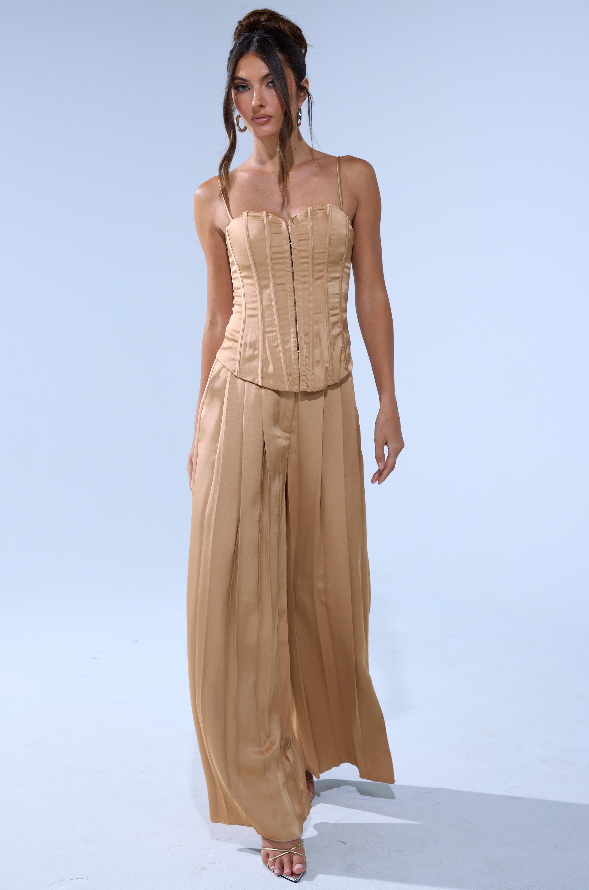 A woman wears the HANSEL MOCHA SATIN CORSET with wide-leg pants, standing against a plain background, her hair styled up with loose strands framing her face.