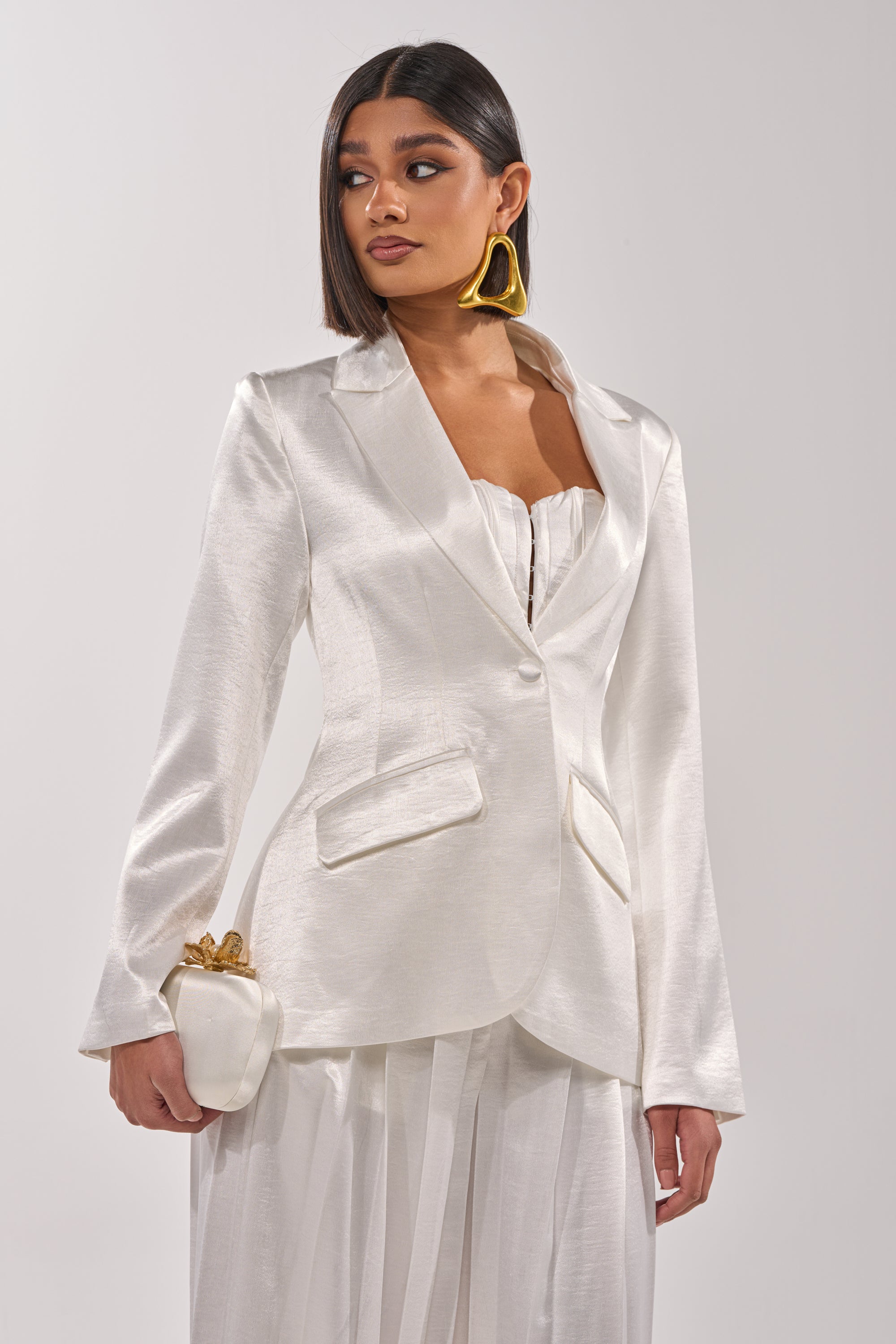 A woman wears the IRIS SATIN BLAZER in white over a matching strapless top and wide-leg trousers, accessorized with a white clutch with gold accents, straight dark hair, and large triangular gold earrings. 2