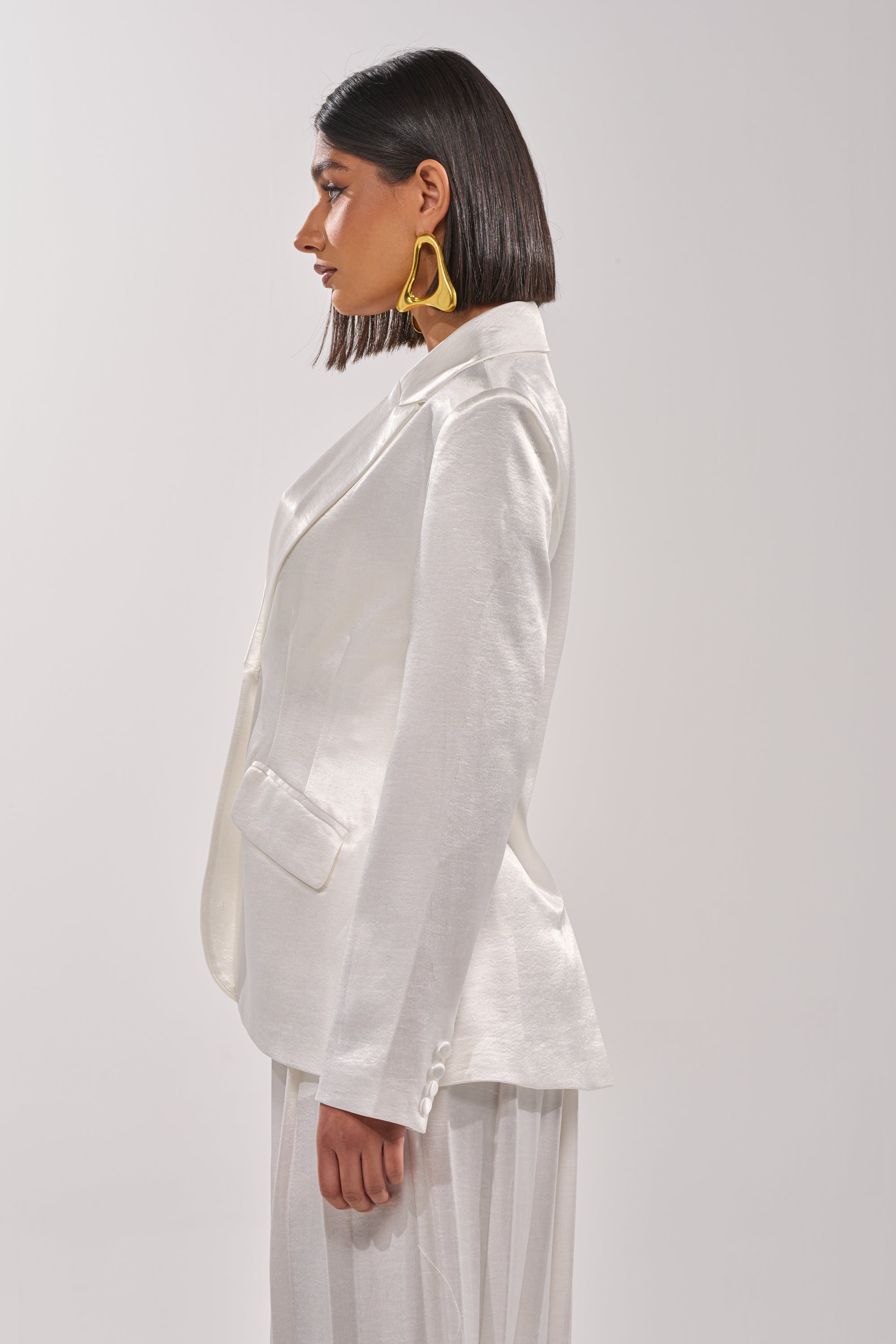A woman with a sleek bob wears the IRIS SATIN BLAZER and wide-leg pants, standing in profile. She accessorizes with large abstract gold earrings against a plain light background. 3