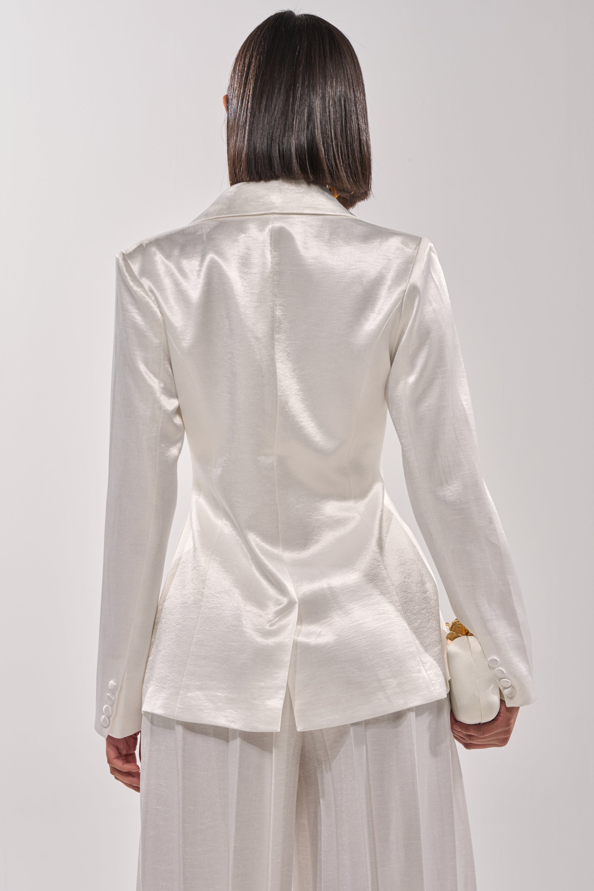 A person with straight, shoulder-length dark hair faces away, wearing the IRIS SATIN BLAZER with a tailored waist and matching wide-leg pants, holding a white clutch against a plain light background. 4