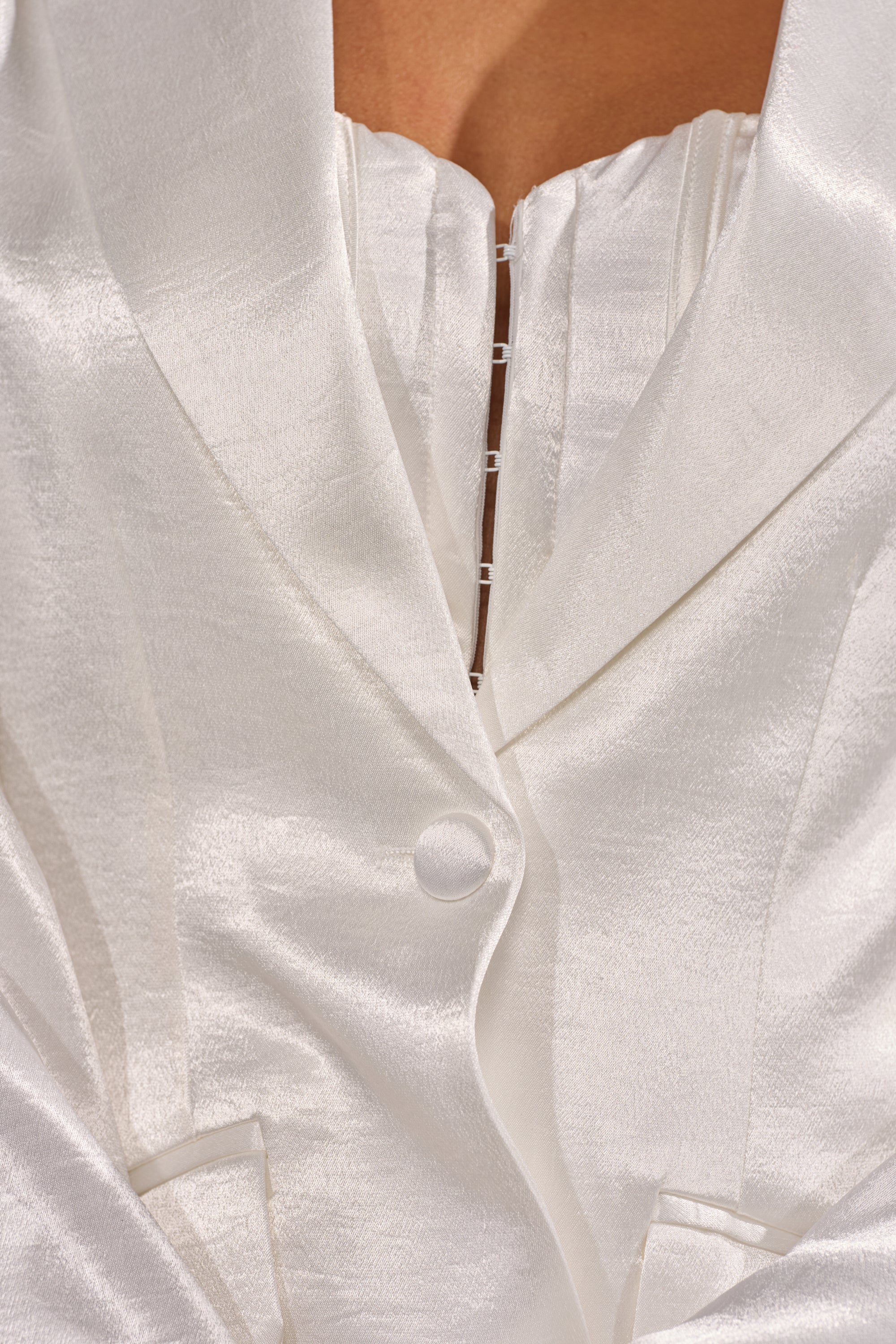 Close-up of a person wearing the IRIS SATIN BLAZER—a white, high-shine satin piece with tailored waist, silver zipper, matching textured top, and details including upper chest, lapel, and buttons. 8