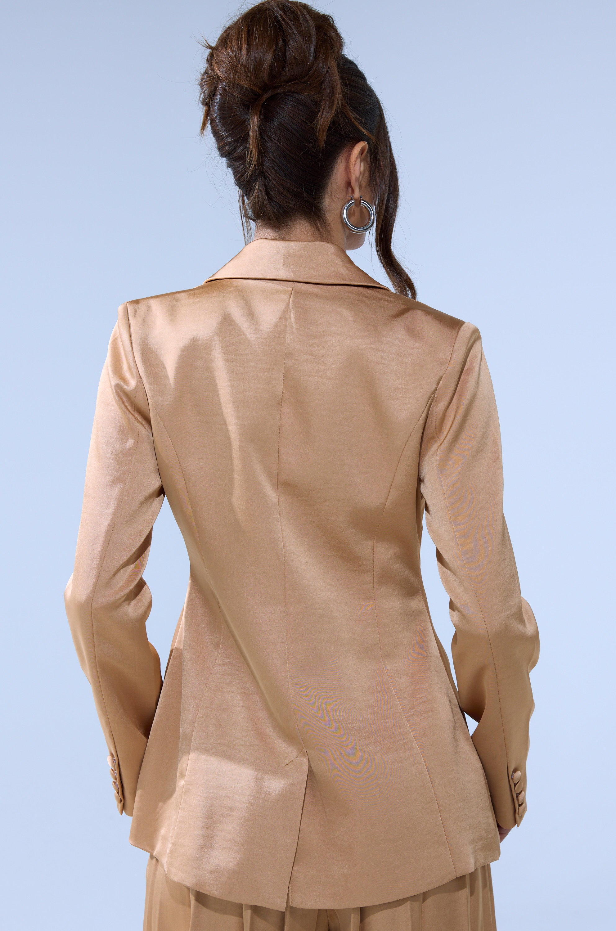 A woman with long brown hair in an updo, wearing the HANSEL MOCHA SATIN BLAZER and hoop earrings, is seen from behind against a light blue background.