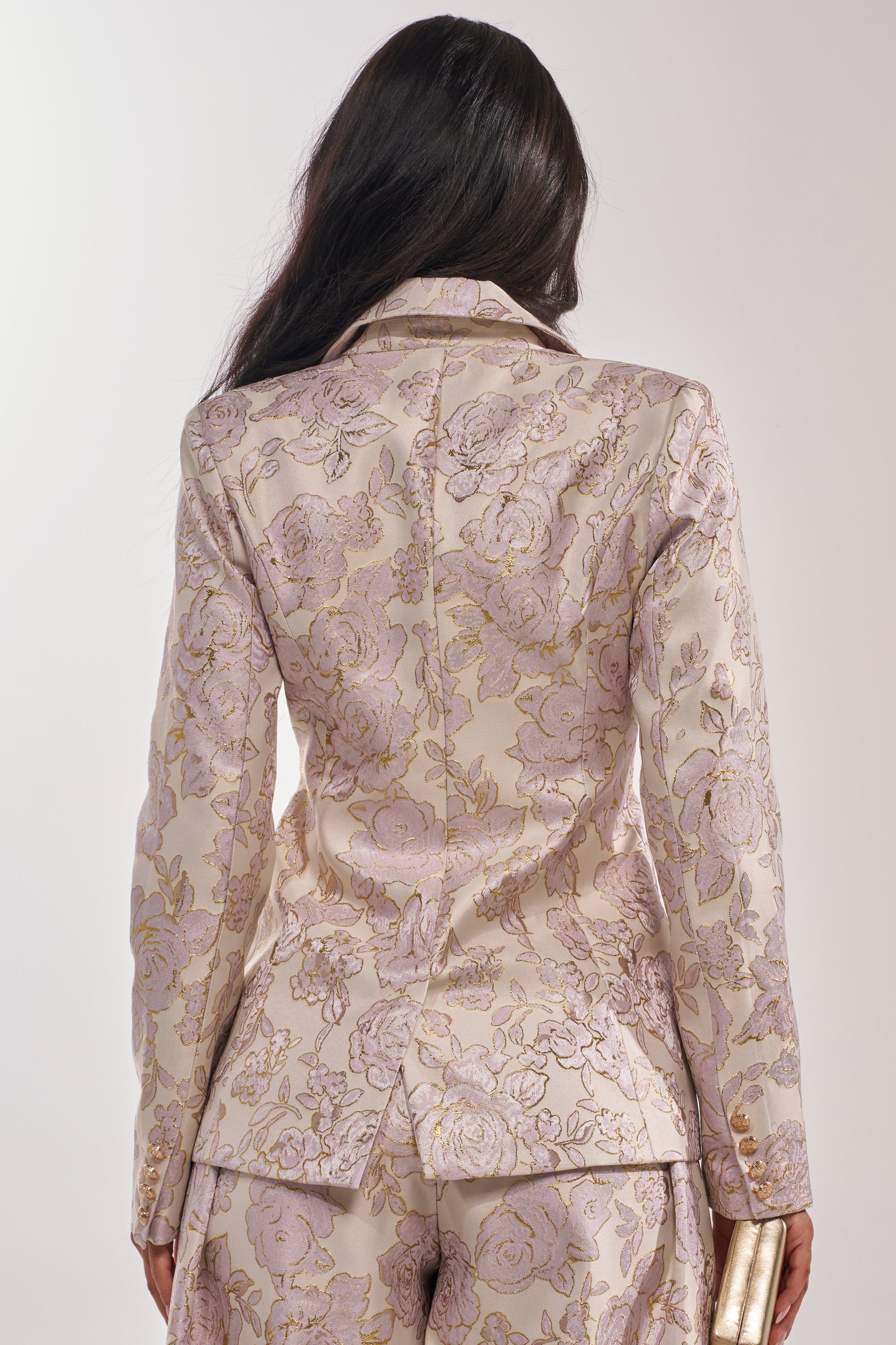 A woman with long dark hair is seen from the back wearing the CHERI BROCADE BLAZER and matching pants in cream and gold floral, holding a small gold clutch in her right hand. 5