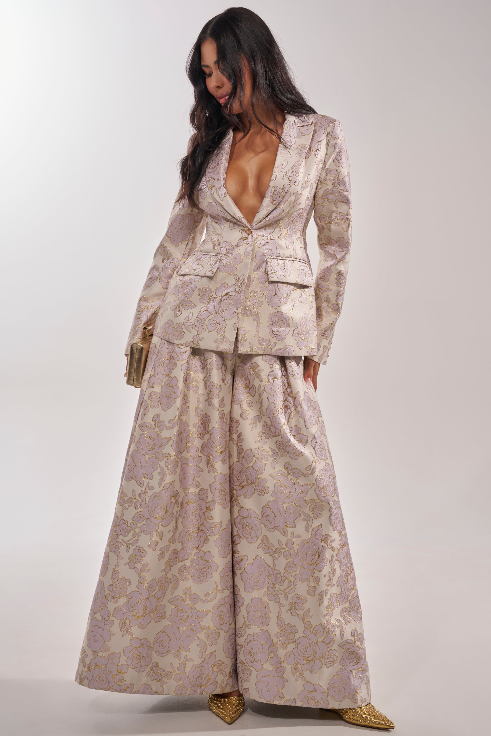 A woman models the CHERI BROCADE BLAZER, a tailored floral piece with a deep V-neck, paired with matching wide-leg, floor-length pants in light gold and lavender, and pointed gold shoes, set against a minimalist background. 6