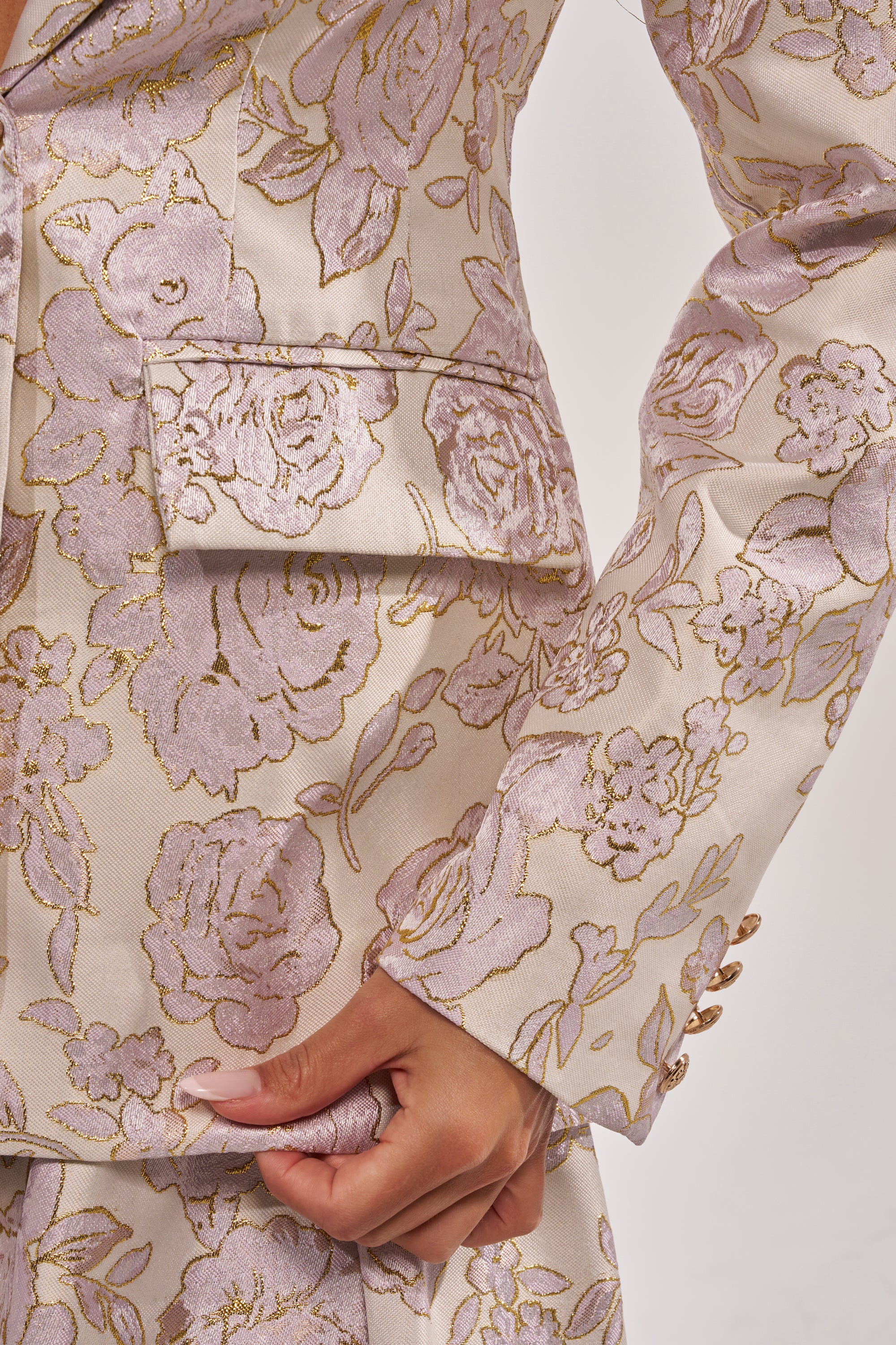 A close-up of a person holding the edge near the pocket of the CHERI BROCADE BLAZER, featuring a cream shade with shiny pink and gold floral patterns. 9
