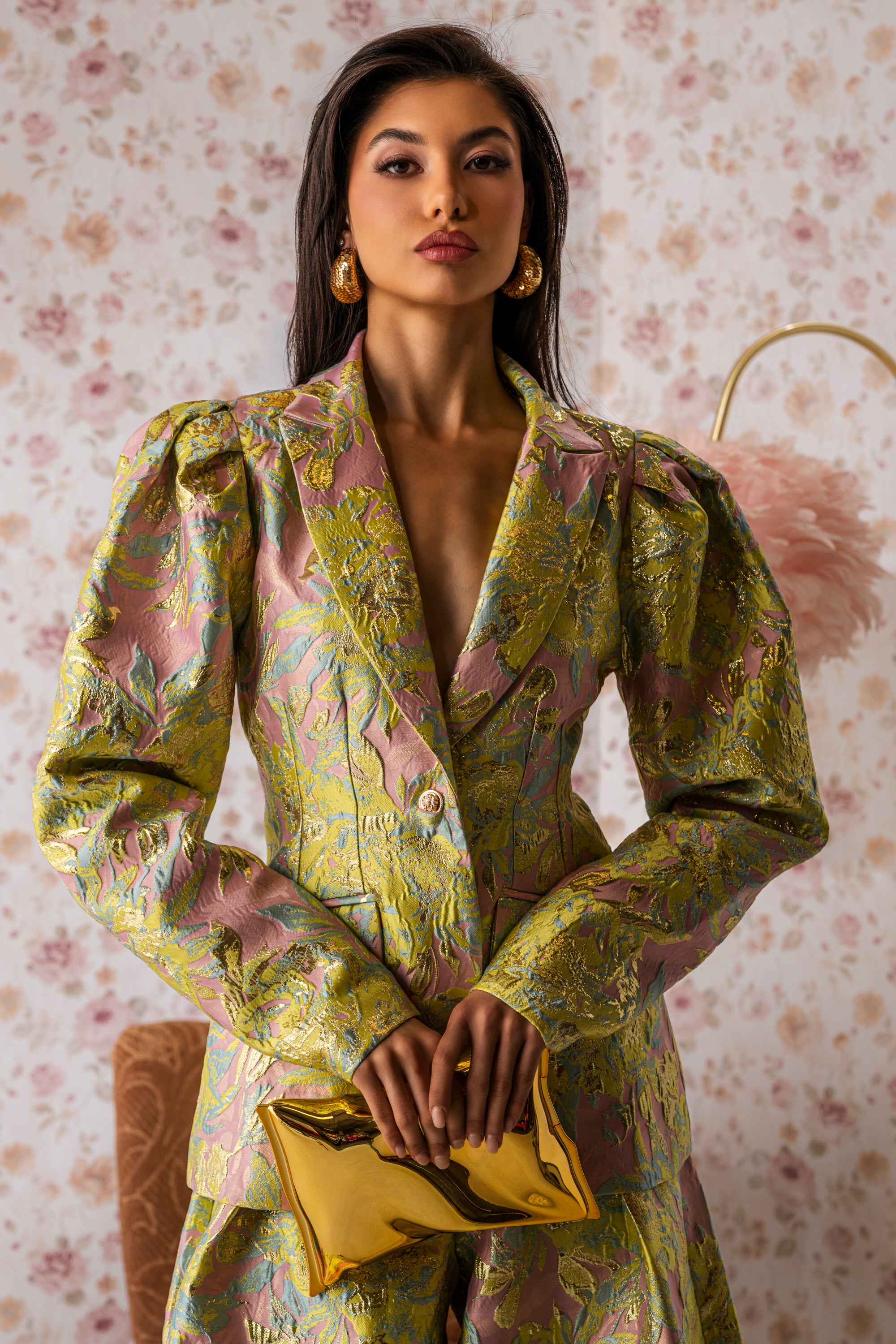 A woman wears the LUCY BROCADE BLAZER in a pastel floral pattern with a jacquard suit, holding a shiny gold clutch and wearing gold earrings. She stands against floral wallpaper, gazing confidently at the camera. 0