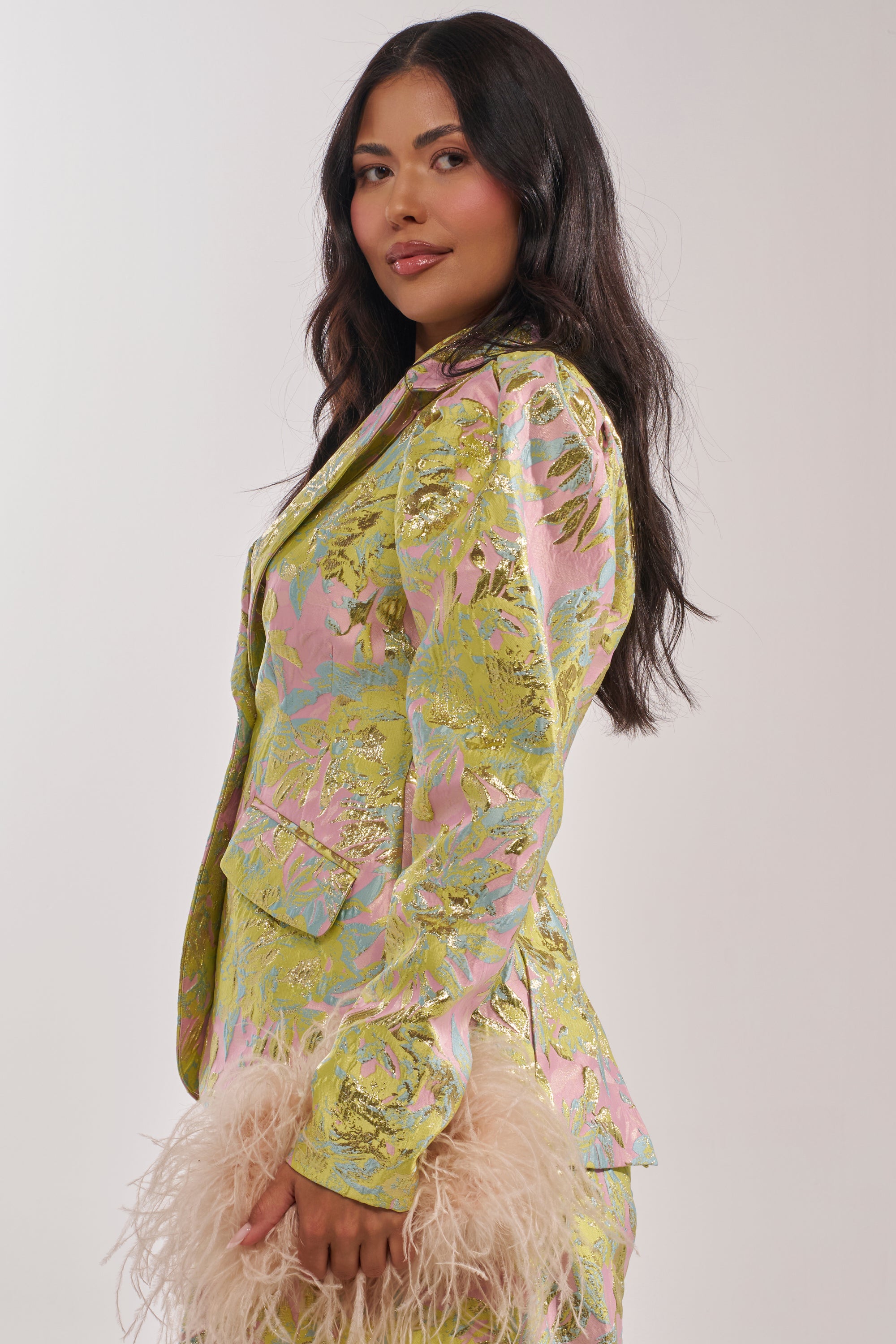 A woman with long dark hair models the LUCY BROCADE BLAZER, featuring a floral-inspired metallic design. She holds a light pink feathered accessory and smiles slightly at the camera against a plain background. 4