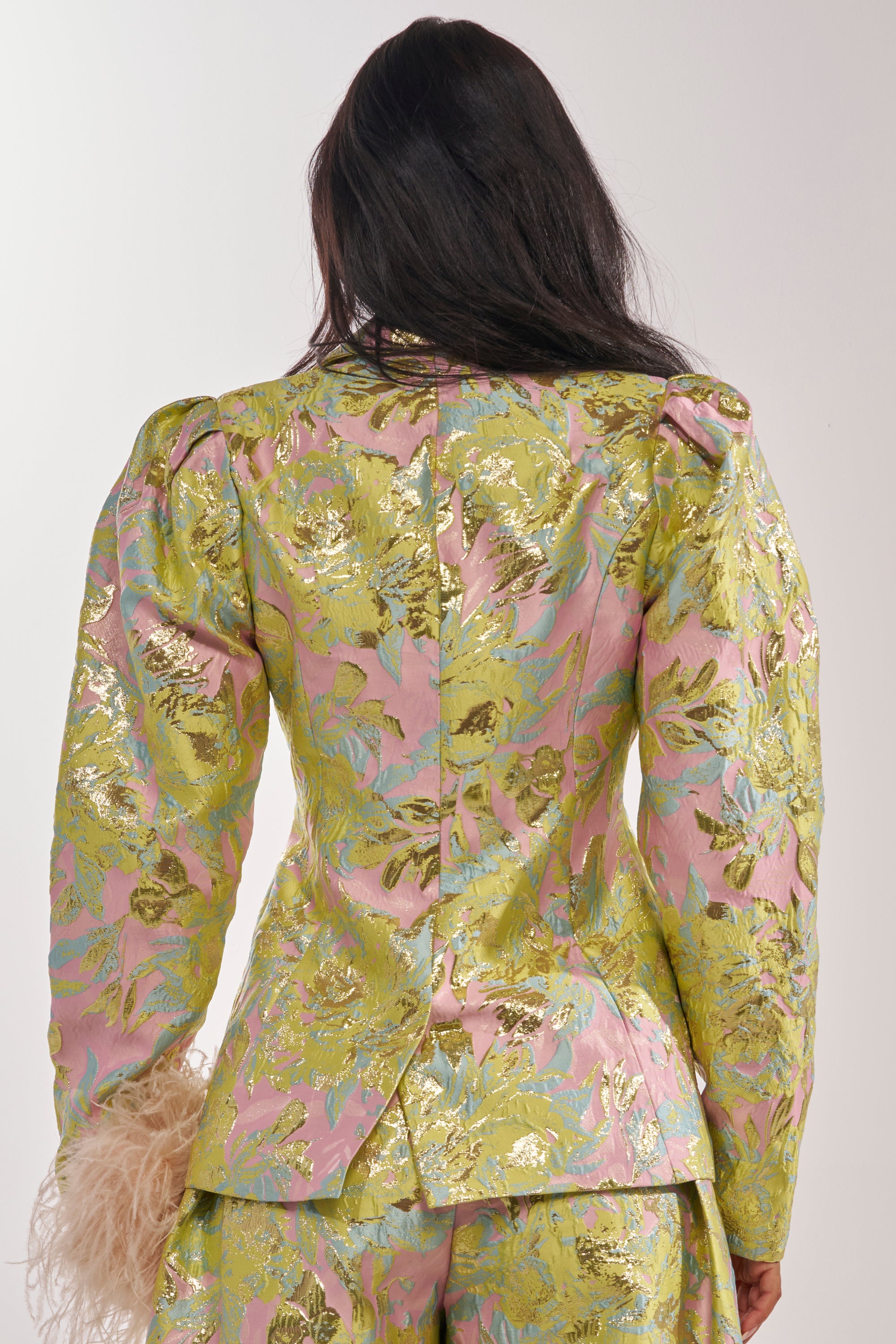 A person with long dark hair is seen from behind, wearing the LUCY BROCADE BLAZER and matching pants in pink metallic brocade with a gold and green floral pattern, puffed shoulders, and holding a beige feathered accessory. 5
