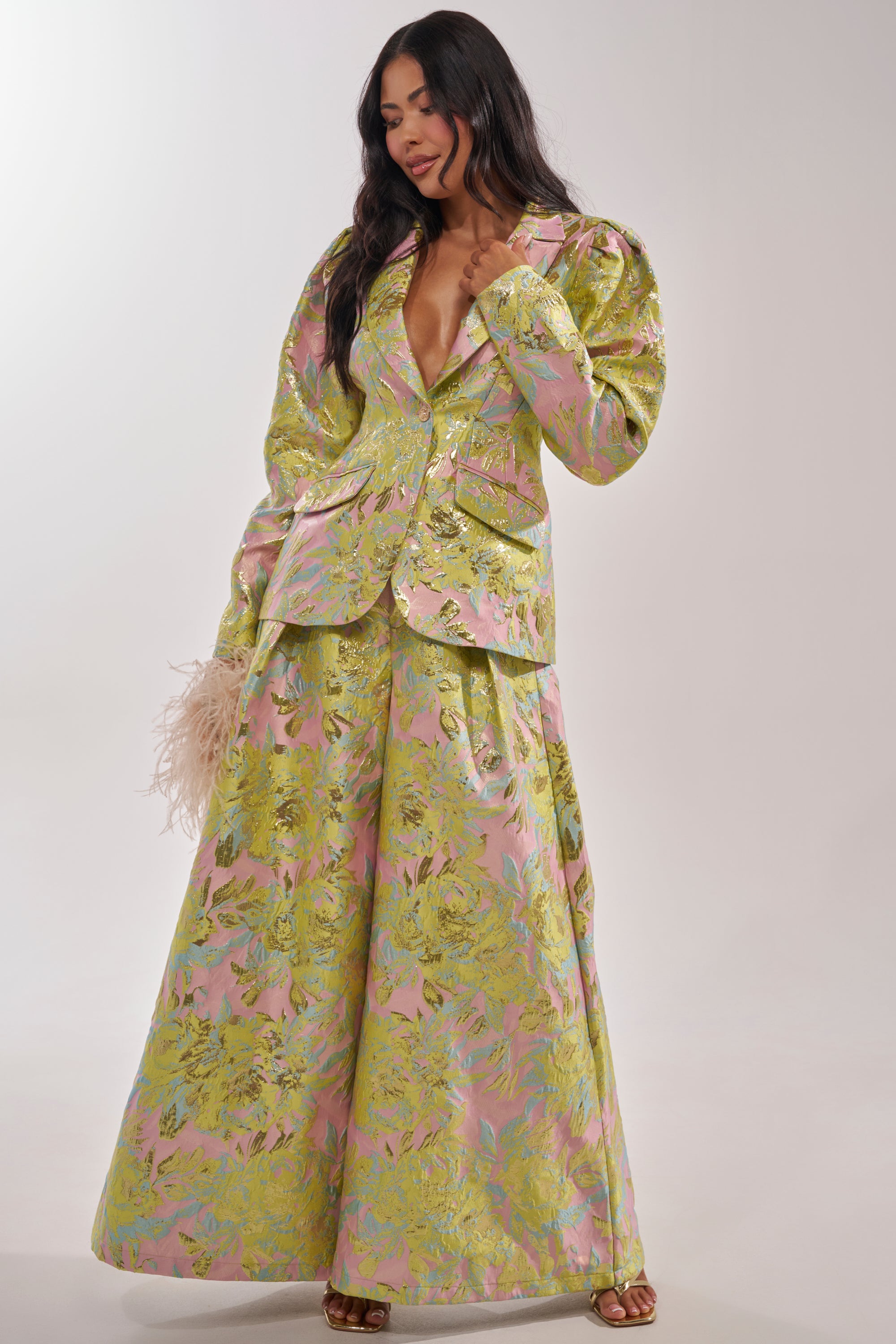 A woman with long hair poses in the LUCY BROCADE BLAZER, a metallic fitted jacket with a pastel green, pink, and gold palette. The blazer features puffed shoulders and is paired with wide-legged pants; she holds a feathered clutch. 7