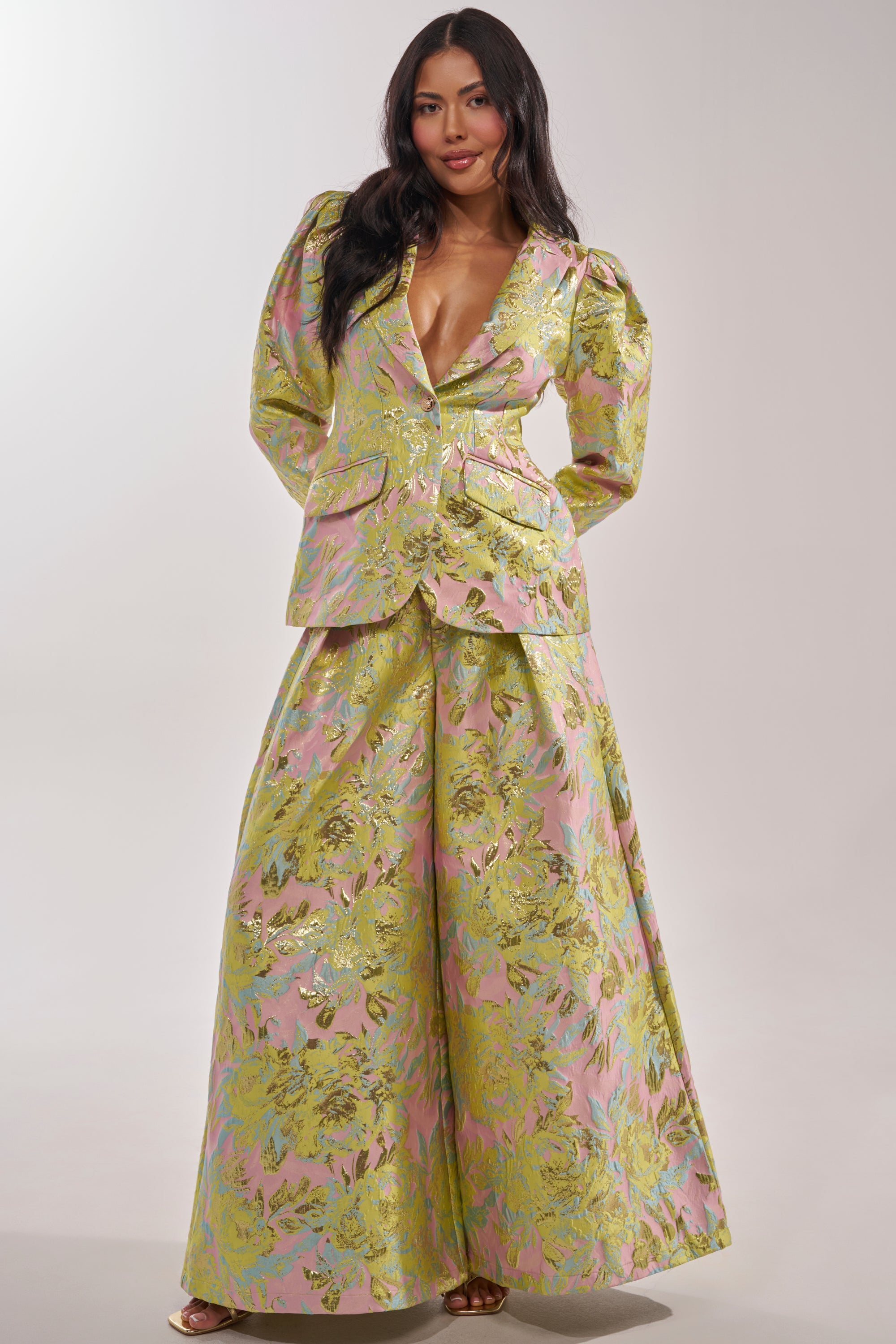 A woman stands confidently in the LUCY BROCADE BLAZER, a pastel metallic suit with wide-leg pants, puffed shoulders, and a deep V-neck, set against a simple light background. 8