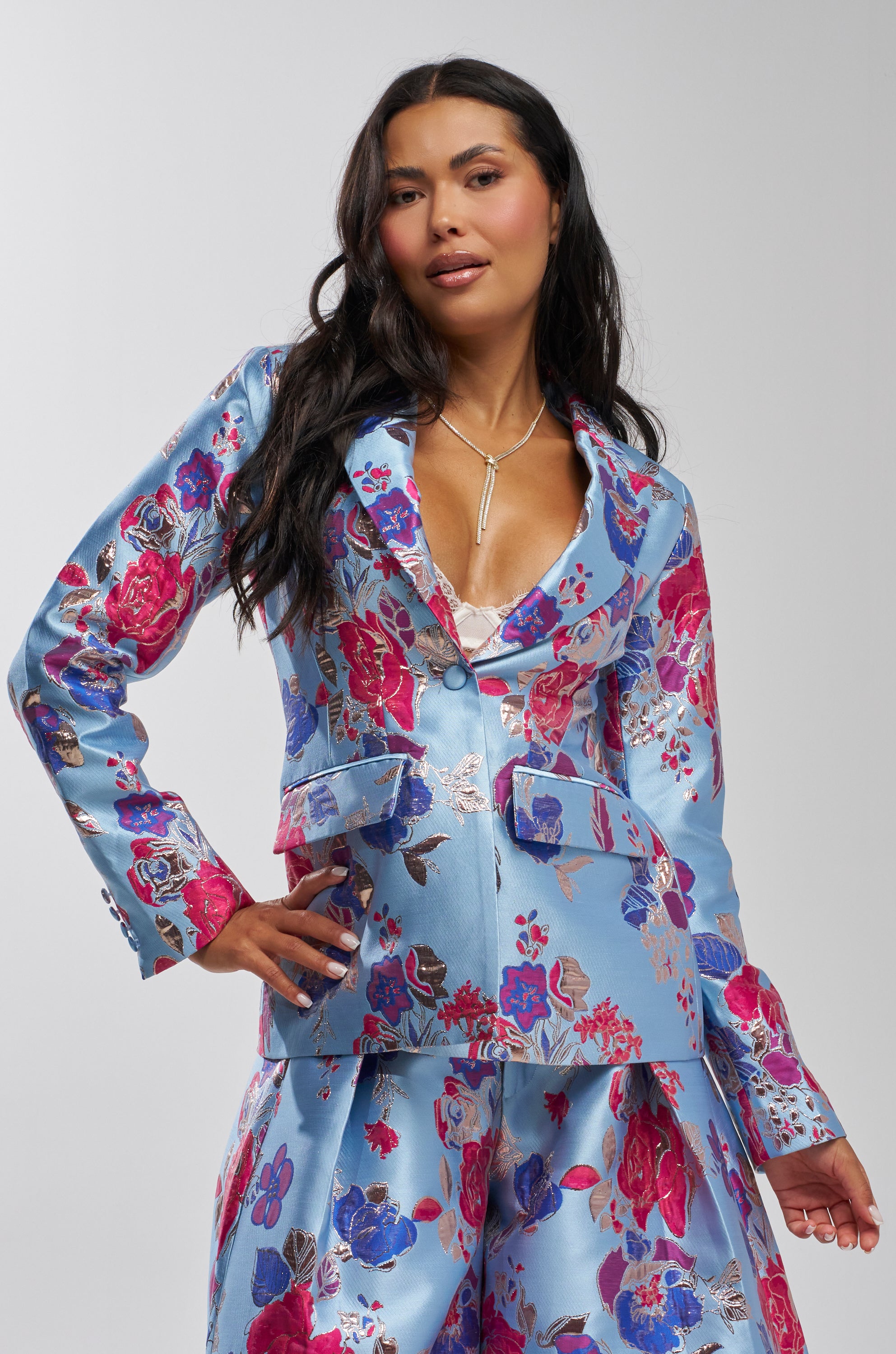 A woman with long dark hair poses confidently with one hand on her hip, wearing the BUY MYSELF FLOWERS BROCADE BLAZER IN BLUE, featuring a shiny light blue floral pattern and bold pink, purple, and silver accents.