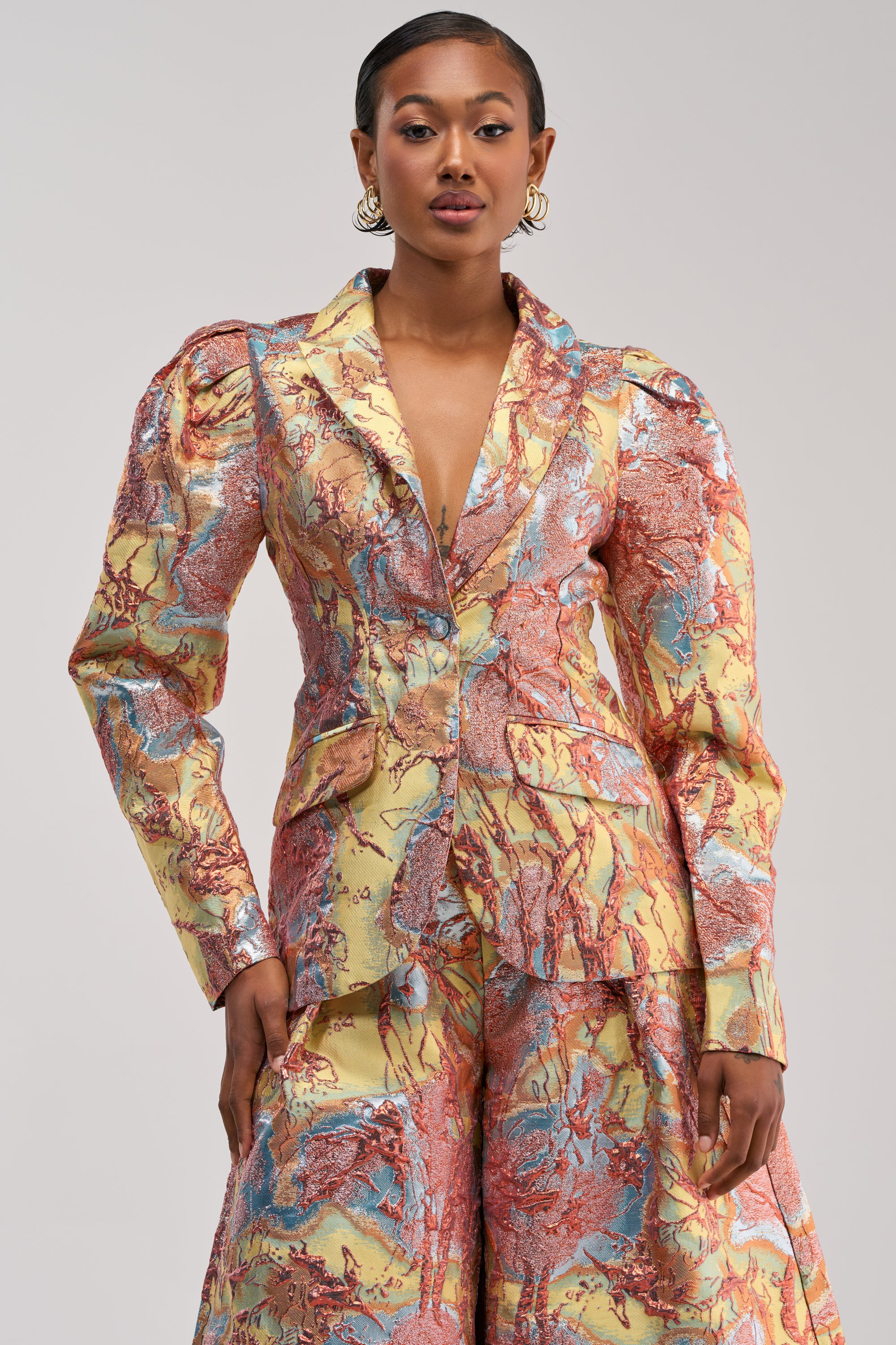 A woman poses in the LUCY BROCADE BLAZER IN BLUE MULTI, a vibrant suit featuring structured shoulders and wide-leg pants with a striking marble pattern in yellow, red, and blue tones. With short, styled hair and gold hoop earrings, she stands against a light background, showcasing this true statement jacket.