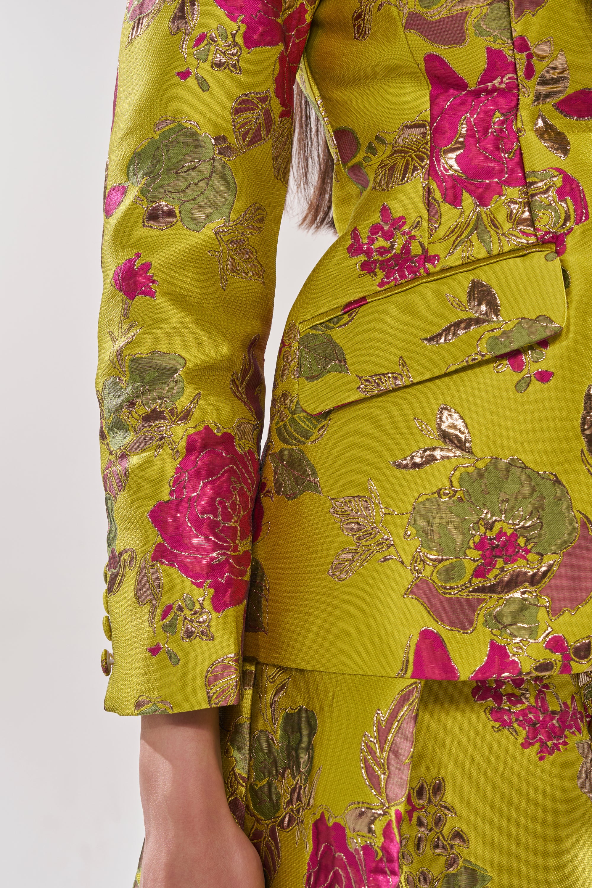 A close-up of a person's arm and torso wearing the BUY MYSELF FLOWERS BROCADE BLAZER IN LIME, featuring bold pink and green floral patterns, metallic embroidery, and richly textured, ornate gold-green brocade fabric.