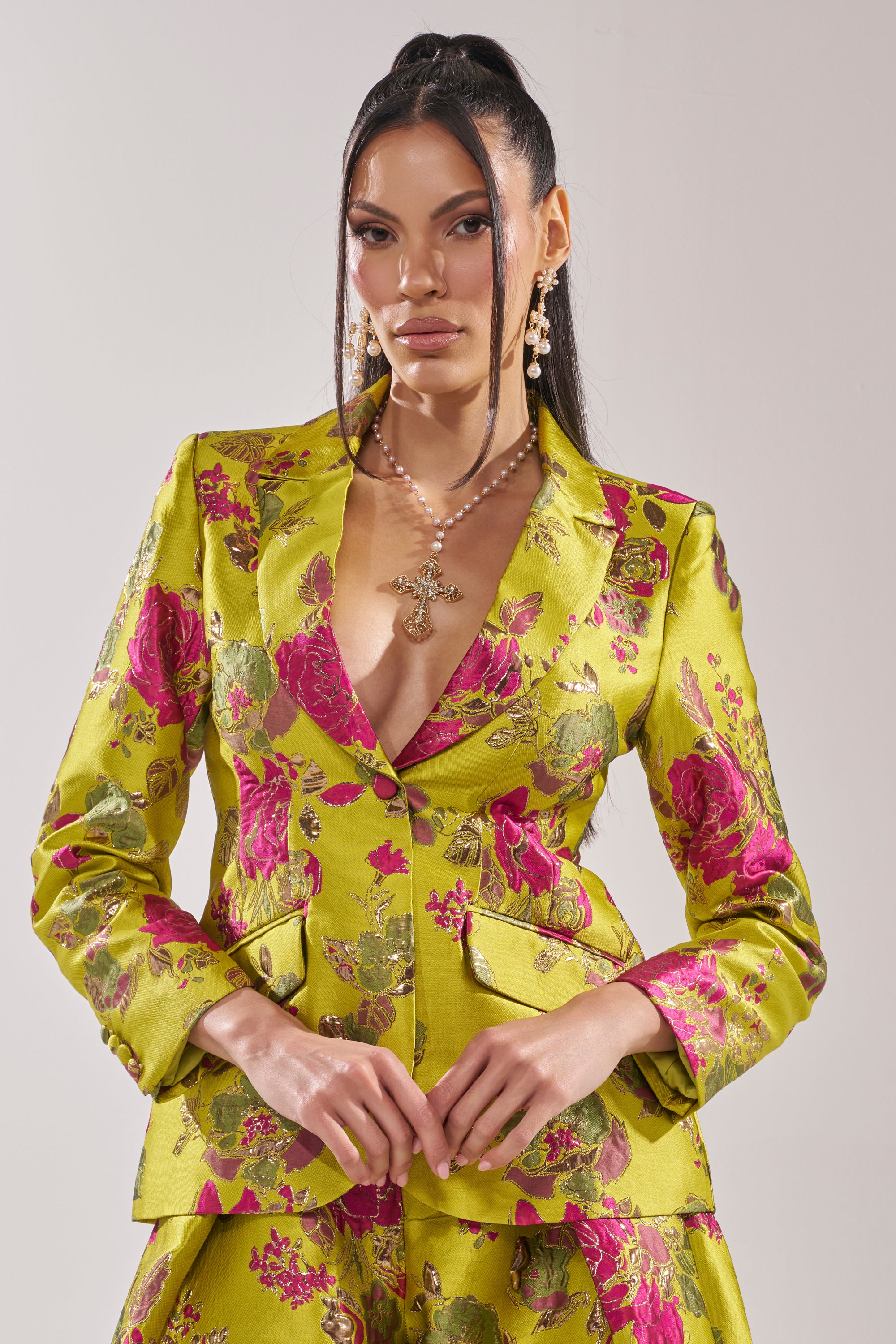 A woman with long dark hair wears the BUY MYSELF FLOWERS BROCADE BLAZER IN LIME, featuring a vivid pink floral pattern. She completes her look with a long necklace and statement earrings against a plain light background.