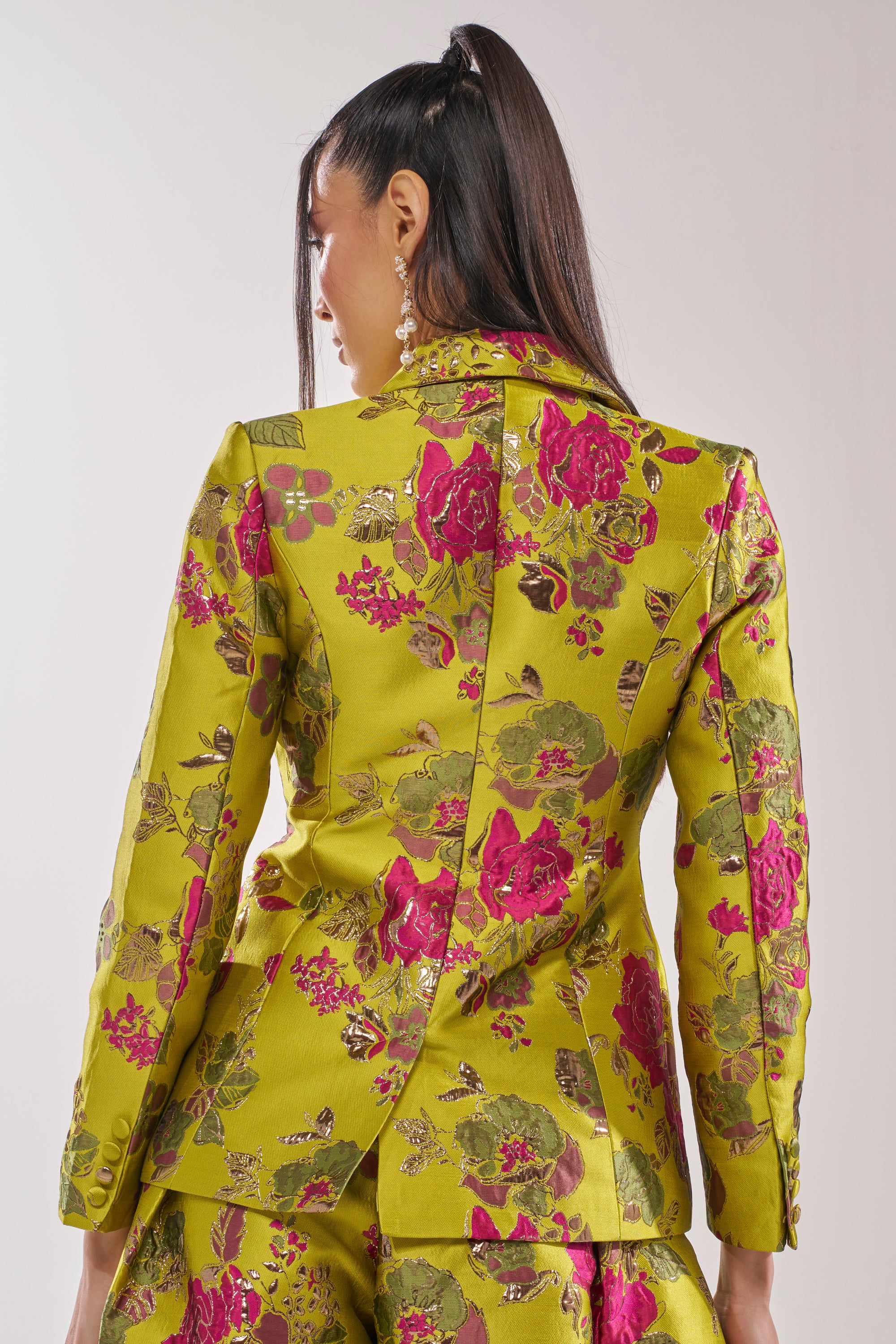 A woman with long dark hair in a high ponytail wears the BUY MYSELF FLOWERS BROCADE BLAZER IN LIME, featuring vibrant pink floral patterns and gold accents, as she faces away to showcase the detailed back of the blazer.