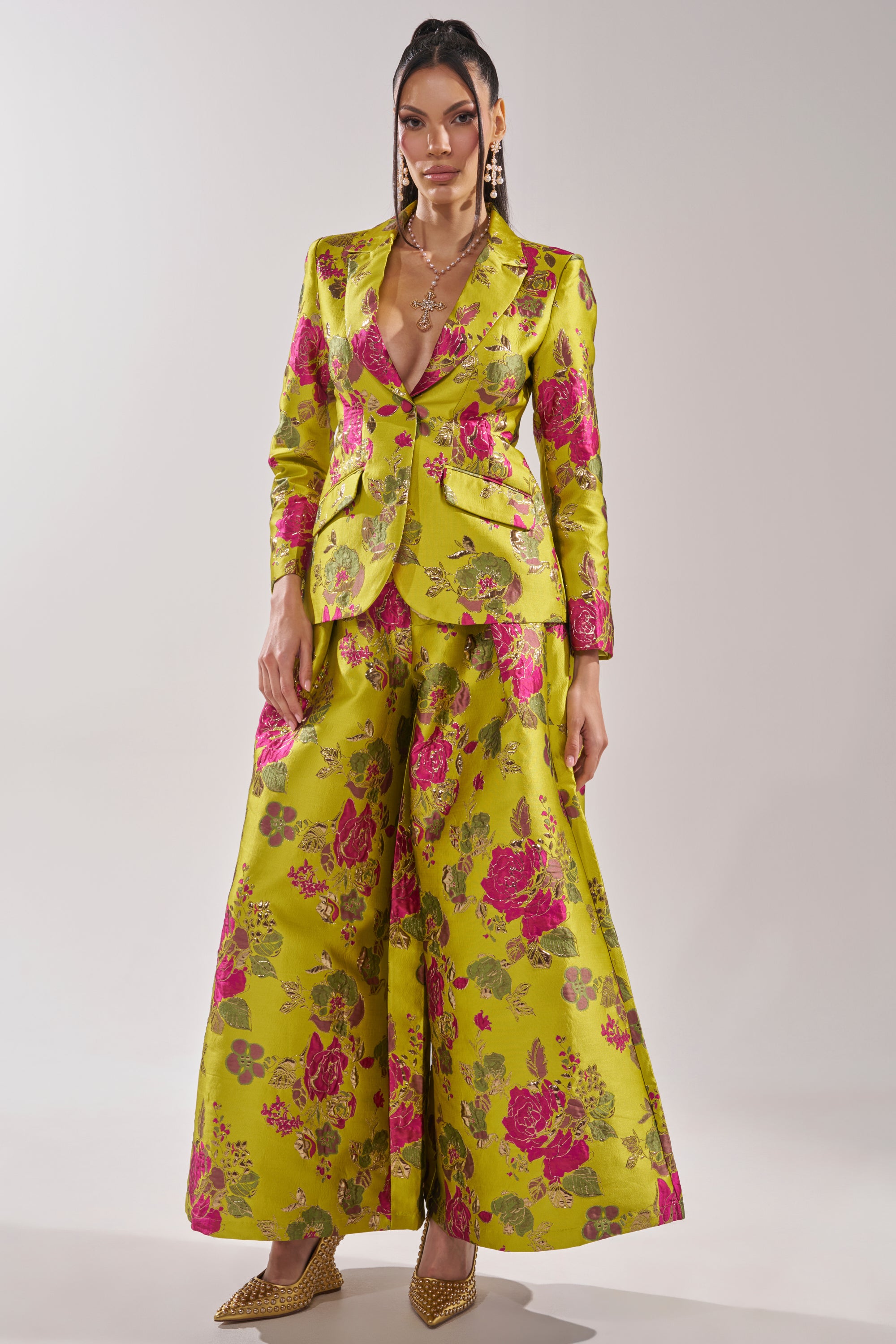A woman poses confidently in the BUY MYSELF FLOWERS BROCADE BLAZER IN LIME—a yellow-green blazer and matching wide-leg trousers with bold pink florals. She accessorizes with gold jewelry and pointed gold heels against a plain background.