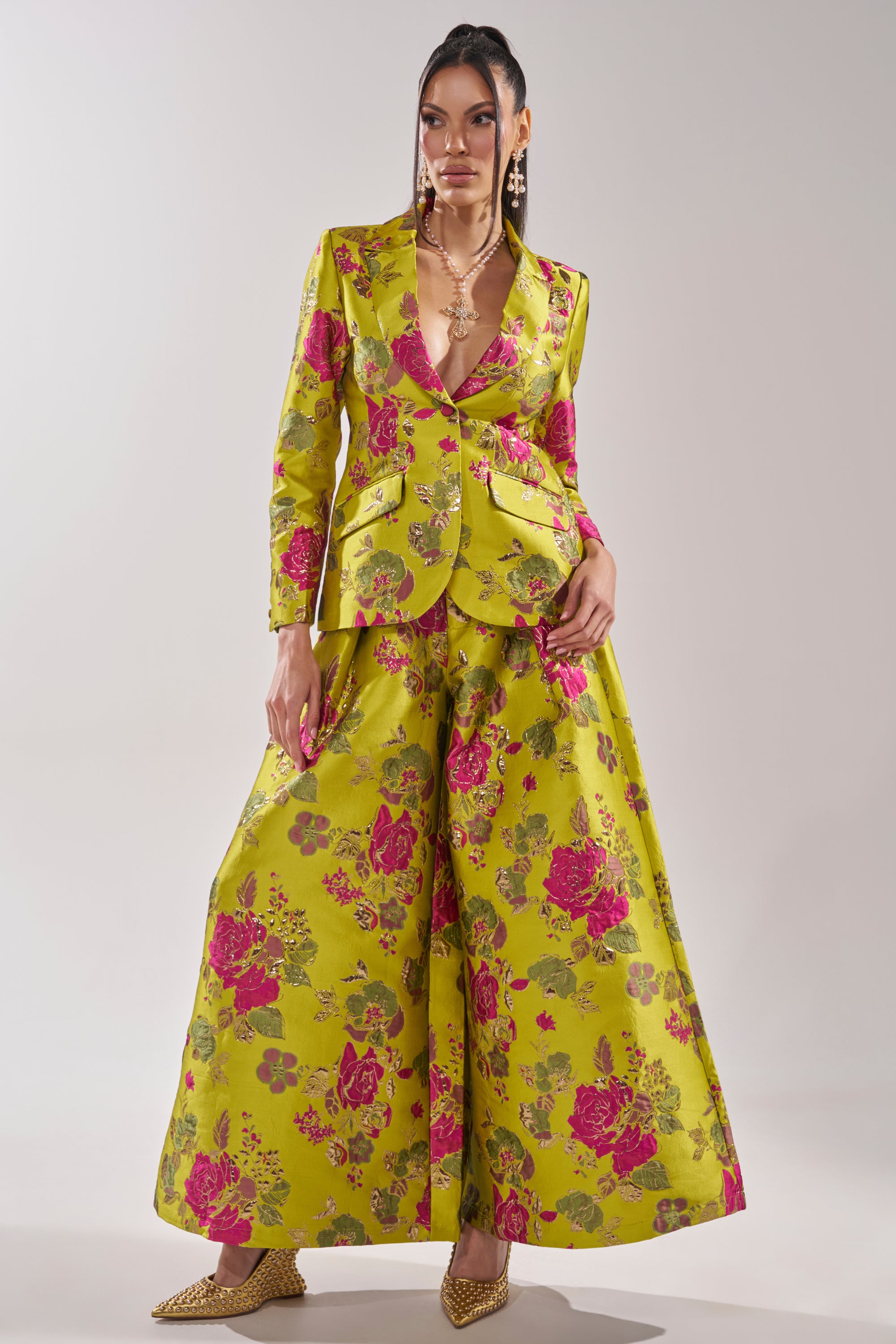 A woman poses in the BUY MYSELF FLOWERS BROCADE BLAZER IN LIME, featuring pink floral patterns, paired with wide-leg trousers. She completes the look with gold pointed heels and layered jewelry against a plain light background.