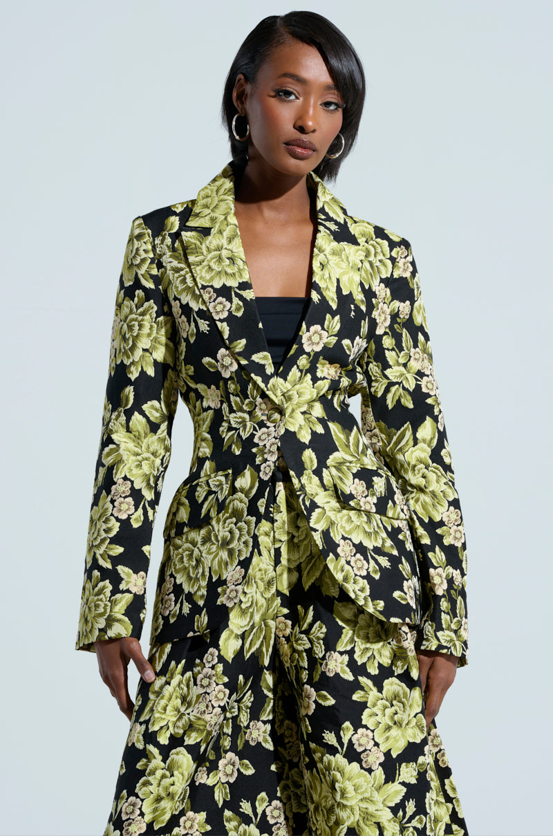A person wearing the BUY MYSELF FLOWERS BROCADE BLAZER paired with matching wide-legged pants, both featuring a chic black and green floral pattern, poses against a light background. They have short, dark hair and complement their look with metallic hoop earrings.