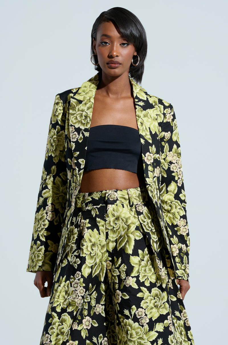 A person wearing the "BUY MYSELF FLOWERS" black and green brocade blazer, embellished with metallic details, and matching wide-legged pants stands against a plain background. They complement the outfit with a strapless black top and hoop earrings, while their hair is styled straight.