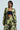 A person wearing the "BUY MYSELF FLOWERS" black and green brocade blazer, embellished with metallic details, and matching wide-legged pants stands against a plain background. They complement the outfit with a strapless black top and hoop earrings, while their hair is styled straight.
