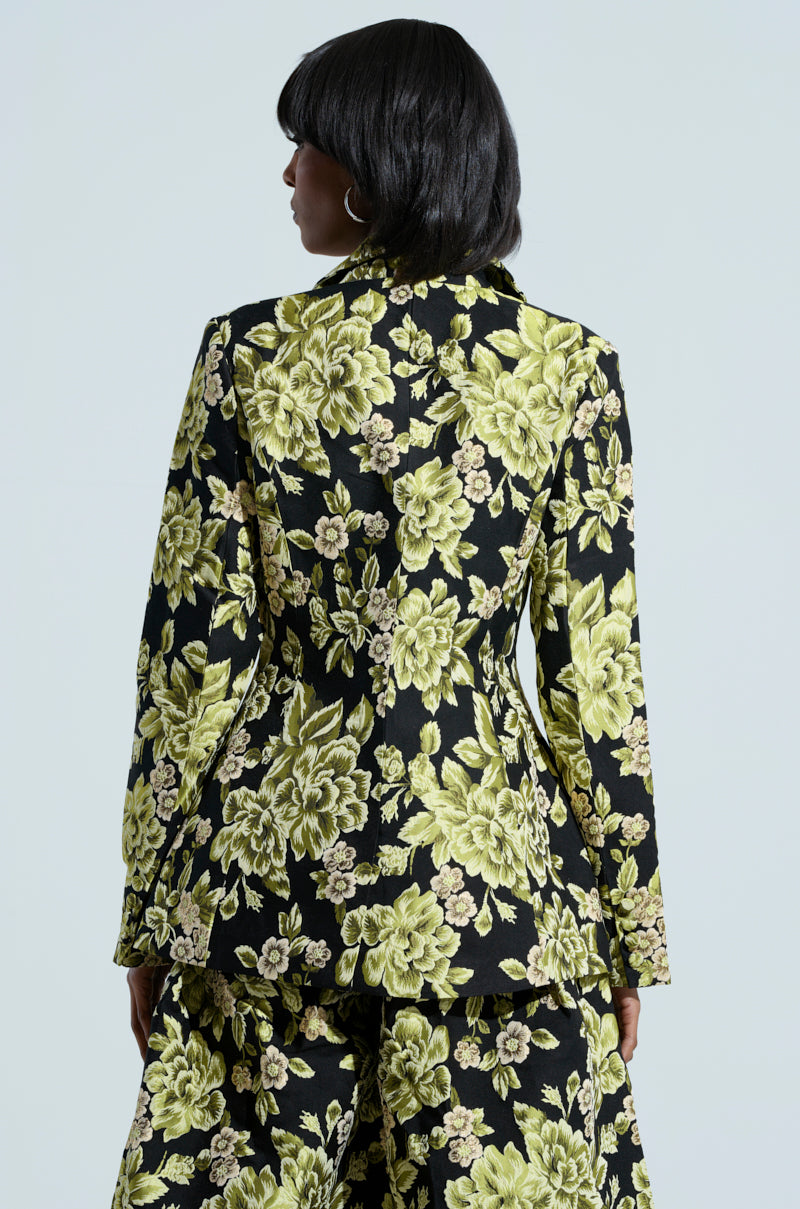 Against a neutral background, the person dons a stylish black suit featuring the BUY MYSELF FLOWERS BROCADE BLAZER, which showcases a striking yellow floral pattern. The ensemble is complemented by wide-leg trousers and adorned with subtle metallic details for an added touch of elegance.