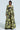 Wearing the BUY MYSELF FLOWERS BROCADE BLAZER, a person stands confidently in a stylish black ensemble adorned with a vibrant green floral pattern. The suit is enhanced with metallic details and a structured design, featuring a long, flowing skirt against a simple light gray background.