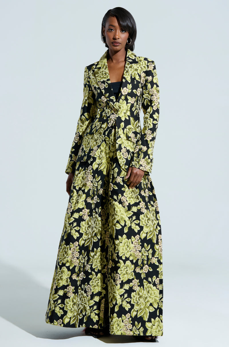 A woman stands confidently wearing the "BUY MYSELF FLOWERS BROCADE BLAZER," a long-sleeved, high-waisted floral suit with a striking black and green pattern. The outfit is completed with wide-legged pants. Her short, sleek black hair contrasts against the simple light background.