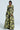 A woman stands confidently wearing the "BUY MYSELF FLOWERS BROCADE BLAZER," a long-sleeved, high-waisted floral suit with a striking black and green pattern. The outfit is completed with wide-legged pants. Her short, sleek black hair contrasts against the simple light background.