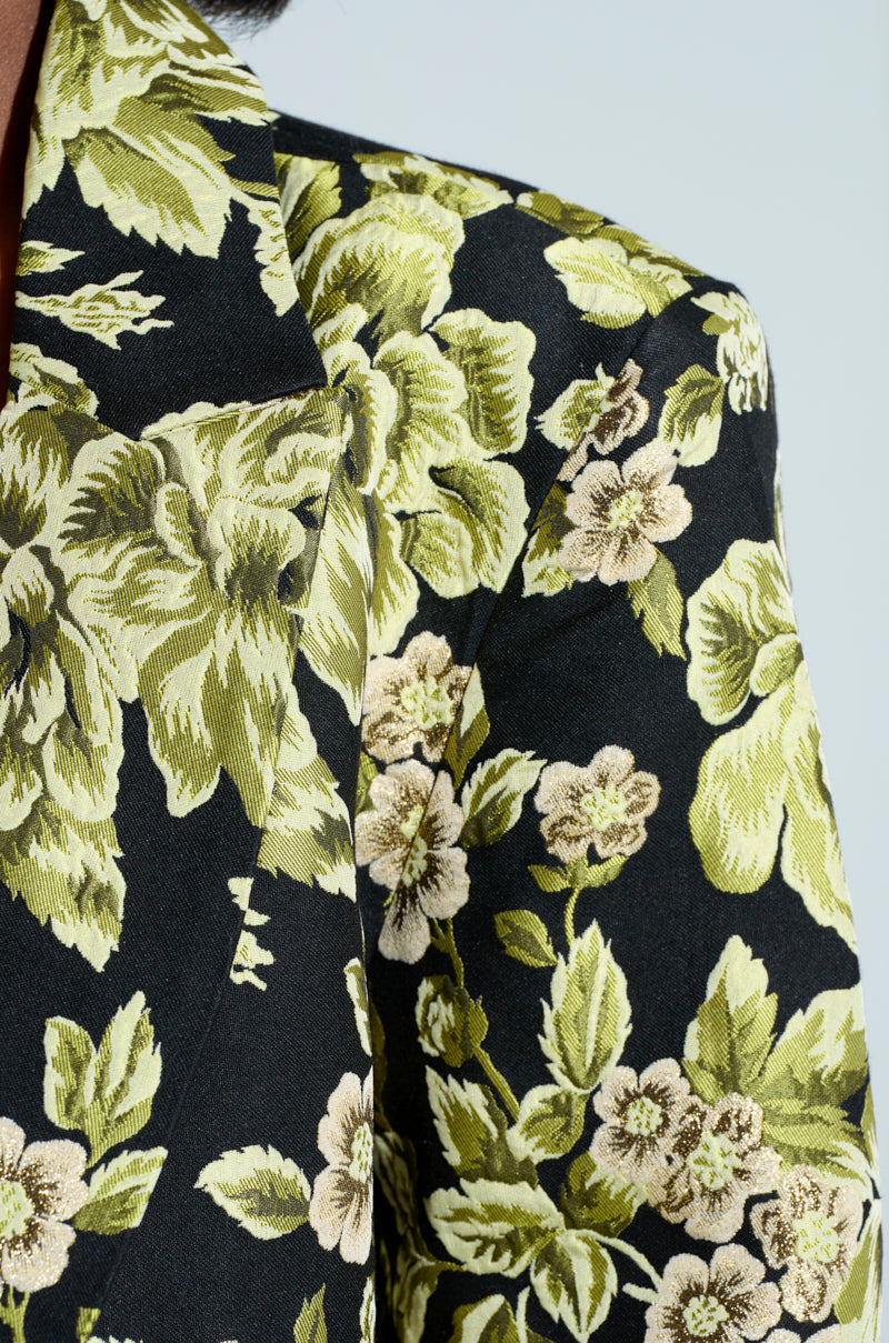 Close-up of a person's shoulder wearing the BUY MYSELF FLOWERS Brocade Blazer, featuring an intricate floral pattern in shades of green and beige with leaves and blossoming flowers, subtly enhanced by metallic details woven into the black fabric.