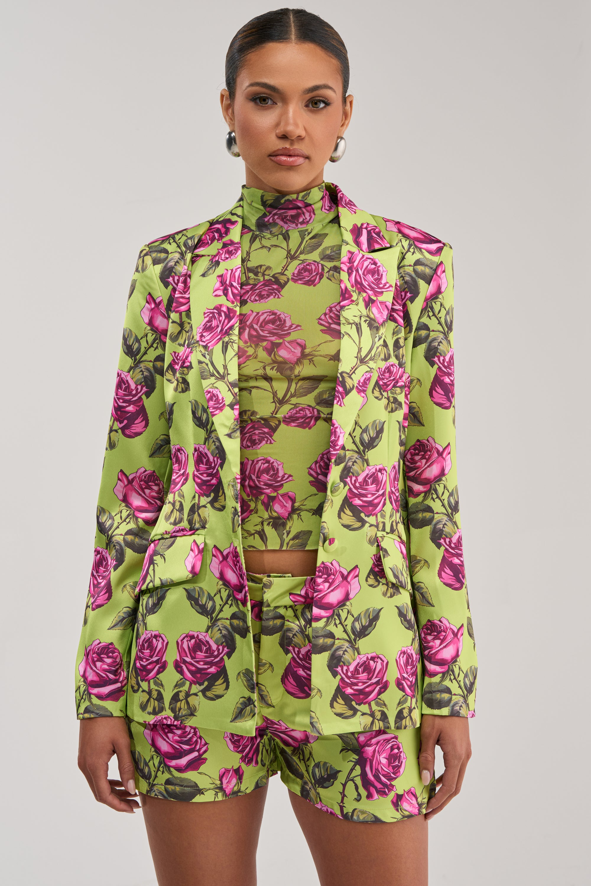 A woman faces forward in the DOZEN ROSES SATIN BLAZER IN CHARTREUSE with matching high-waisted shorts and a sheer floral turtleneck. The plain background highlights this vibrant, formal statement look.