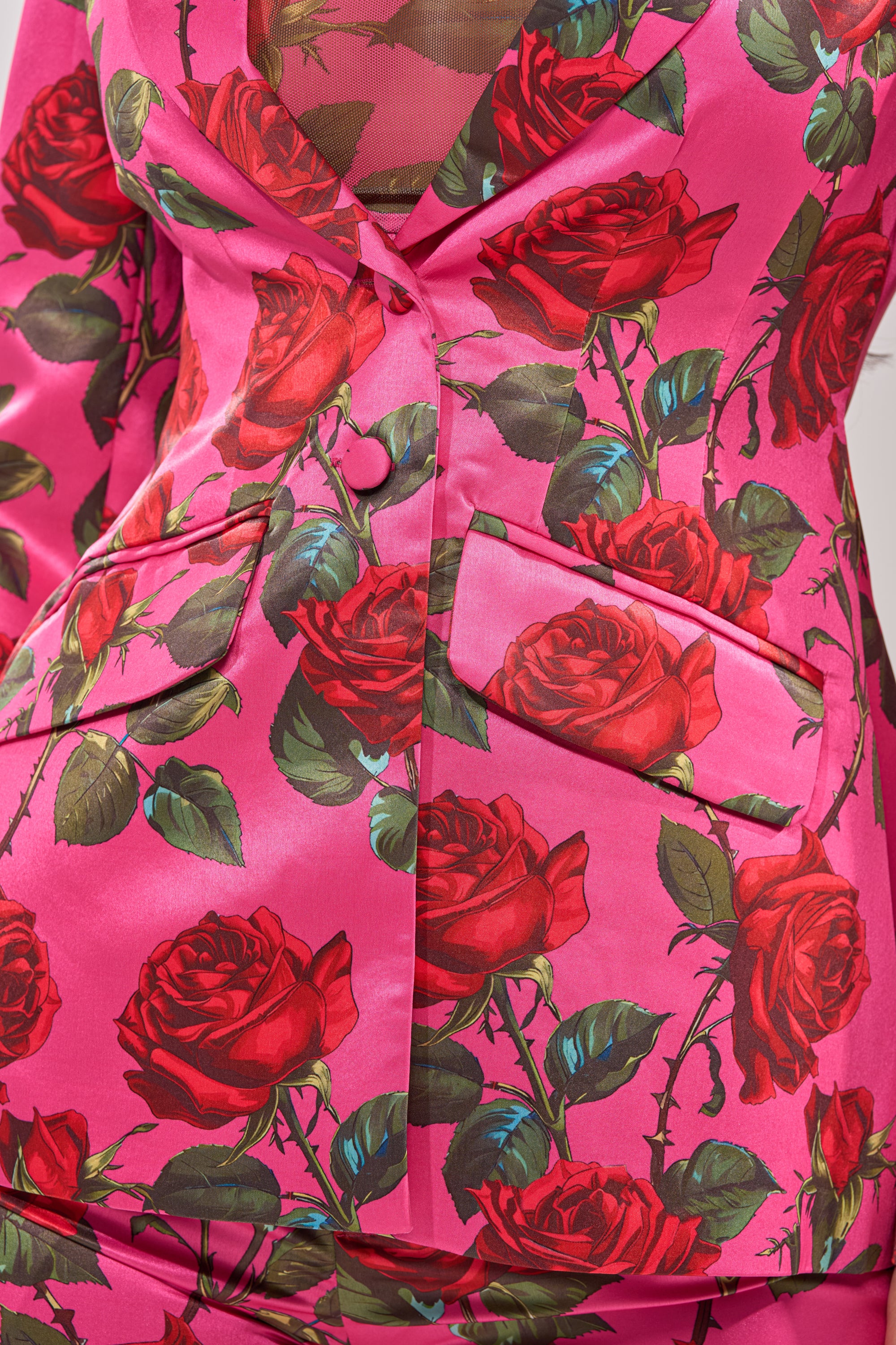 Close-up of the DOZEN ROSES SATIN BLAZER IN FUCHSIA—tailored fit with vivid red rose and green leaf print, two front pockets, and buttons. A bold floral satin piece for standout layering and a striking visual statement. 11