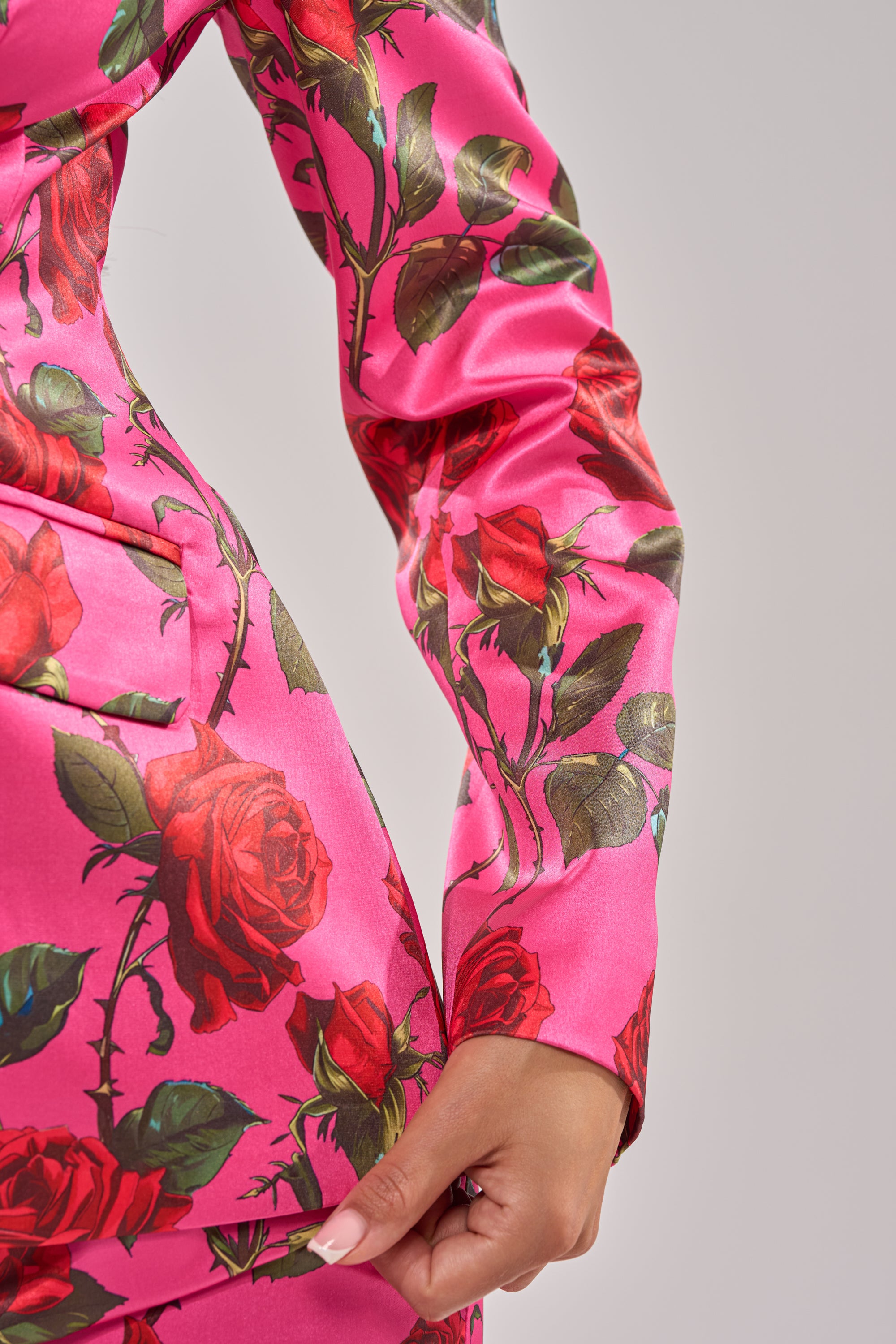 A person models the DOZEN ROSES SATIN BLAZER IN FUCHSIA, featuring a vibrant pink rose-print and tailored fit, shown from waist to mid-arm while gently holding the fabric against a plain light grey background. 12