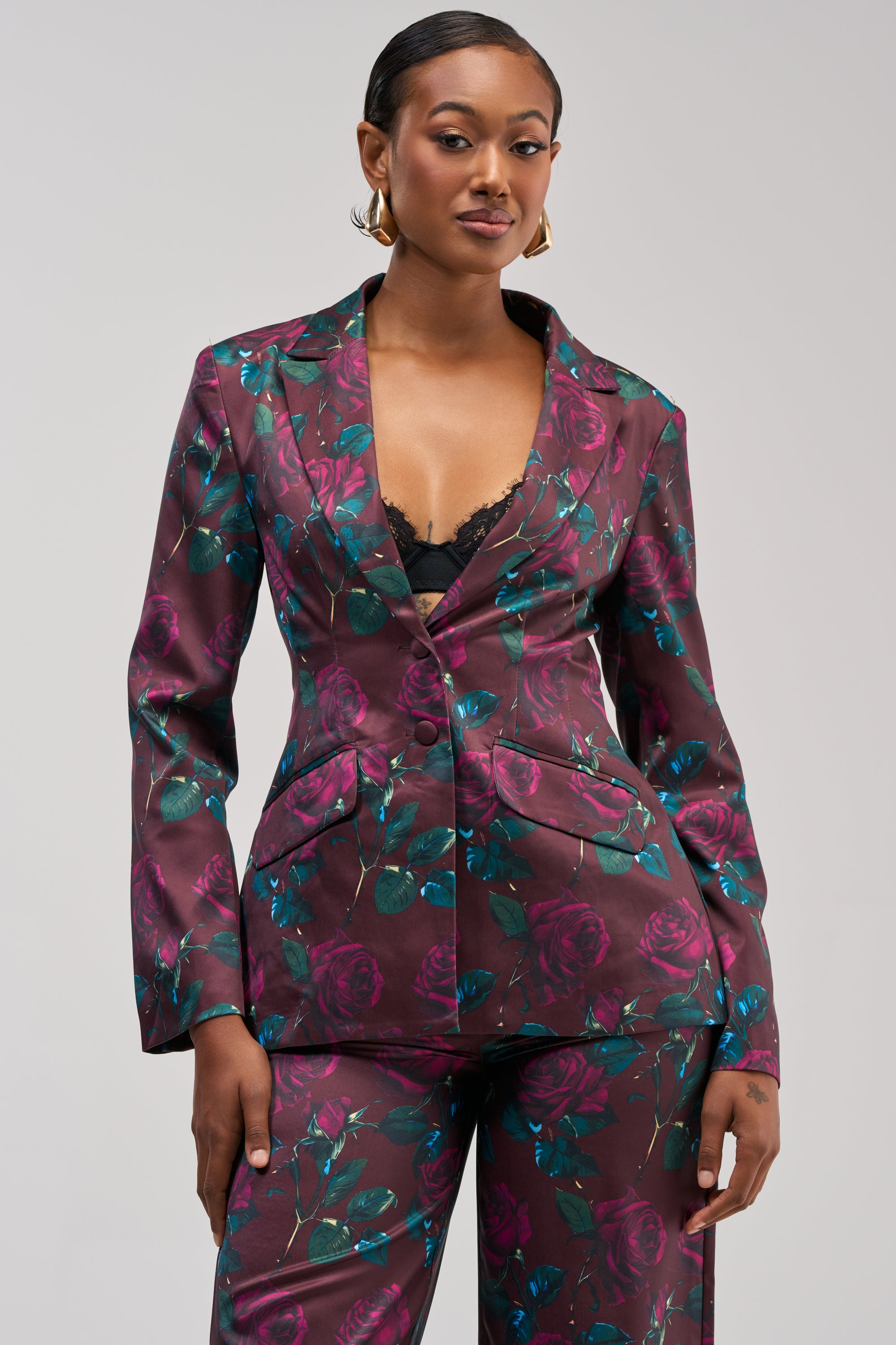 A woman stands confidently against a plain light background, wearing the DOZEN ROSES SATIN BLAZER IN BURGUNDY featuring a rose print, deep neckline, and layered over a lace camisole.