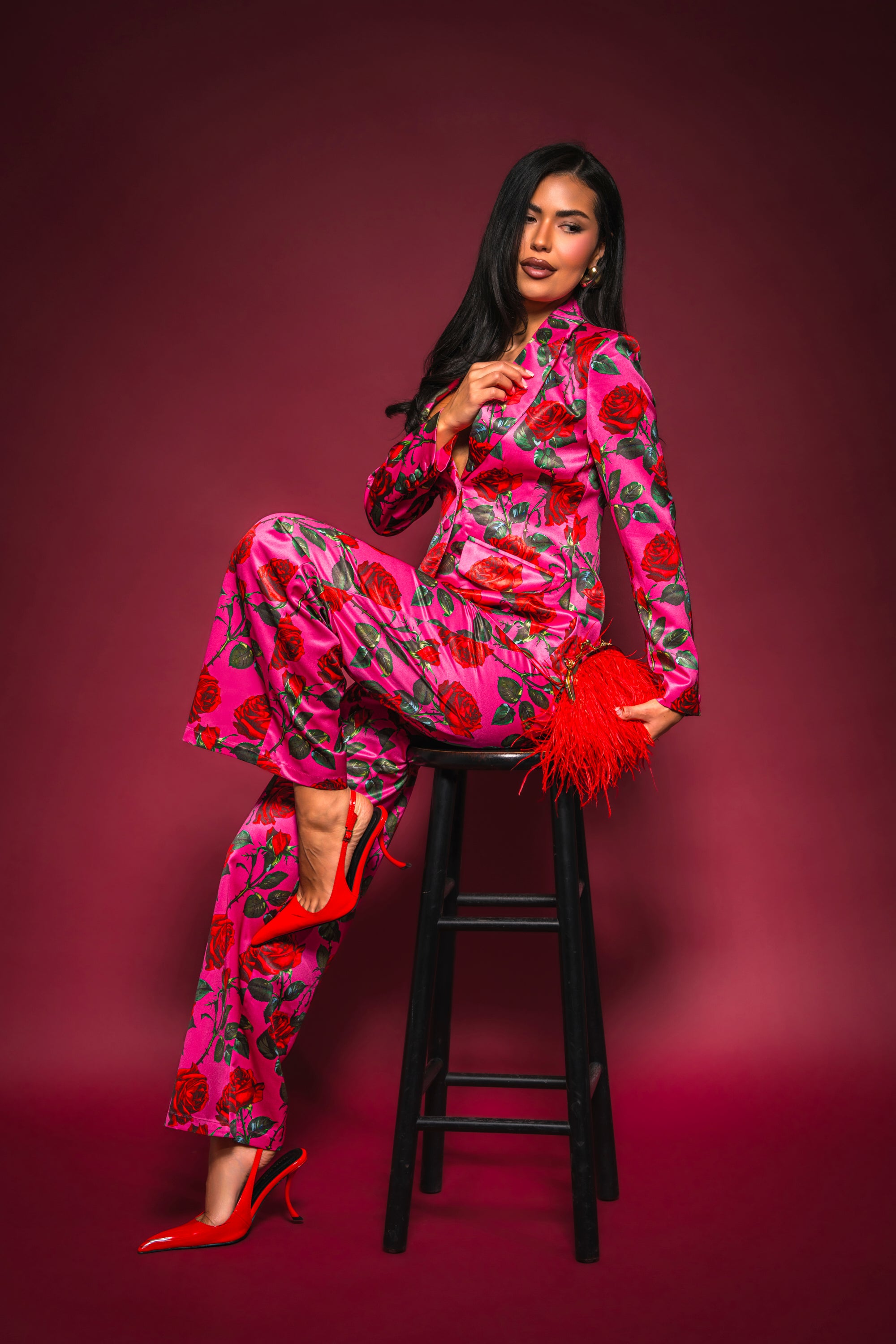 Wearing the DOZEN ROSES SATIN BLAZER IN FUCHSIA, a woman sits confidently on a black stool against a dark pink background, pairing her bold rose-print look with red heels and a feathered red clutch for standout style. 0