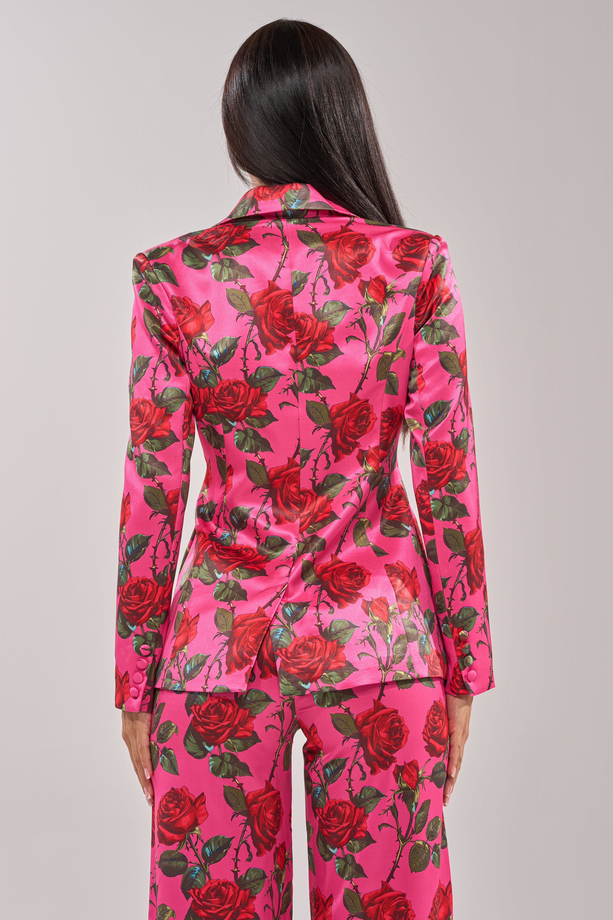 A person with long dark hair is seen from behind, wearing the DOZEN ROSES SATIN BLAZER IN FUCHSIA with bold red rose and green leaf prints, set against a plain light background. 7