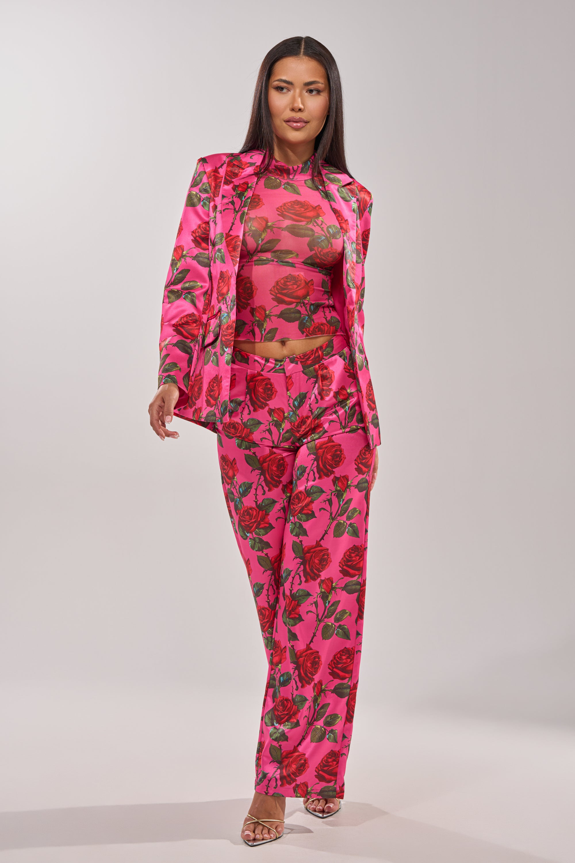 A woman wears the DOZEN ROSES SATIN BLAZER IN FUCHSIA with matching pants and a sheer top. Her long dark hair and confident pose in heels highlight bold, elegant layering against a neutral background. 10