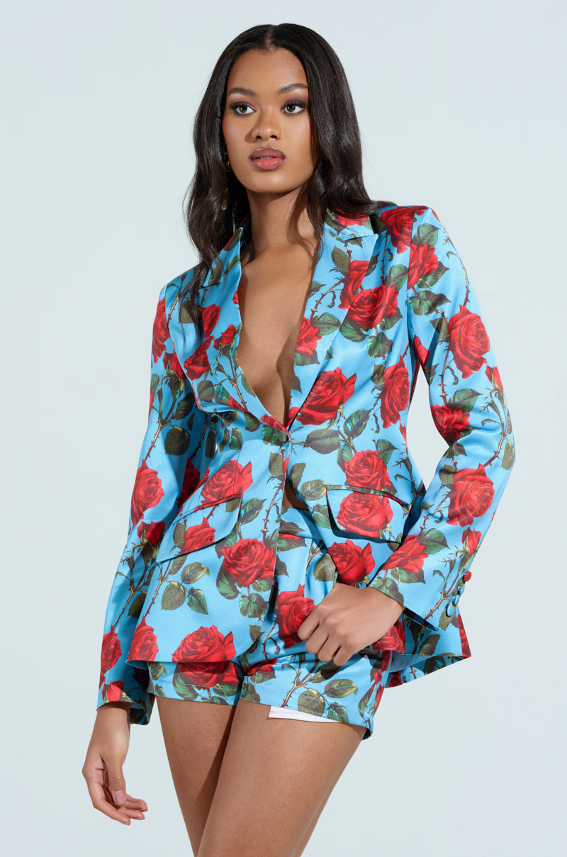 A woman exudes confidence in a vibrant blue DOZEN ROSES SATIN BLAZER paired with matching red-patterned shorts. Her dark hair cascades in soft waves, posing gracefully against a plain backdrop to showcase her chic ensemble.