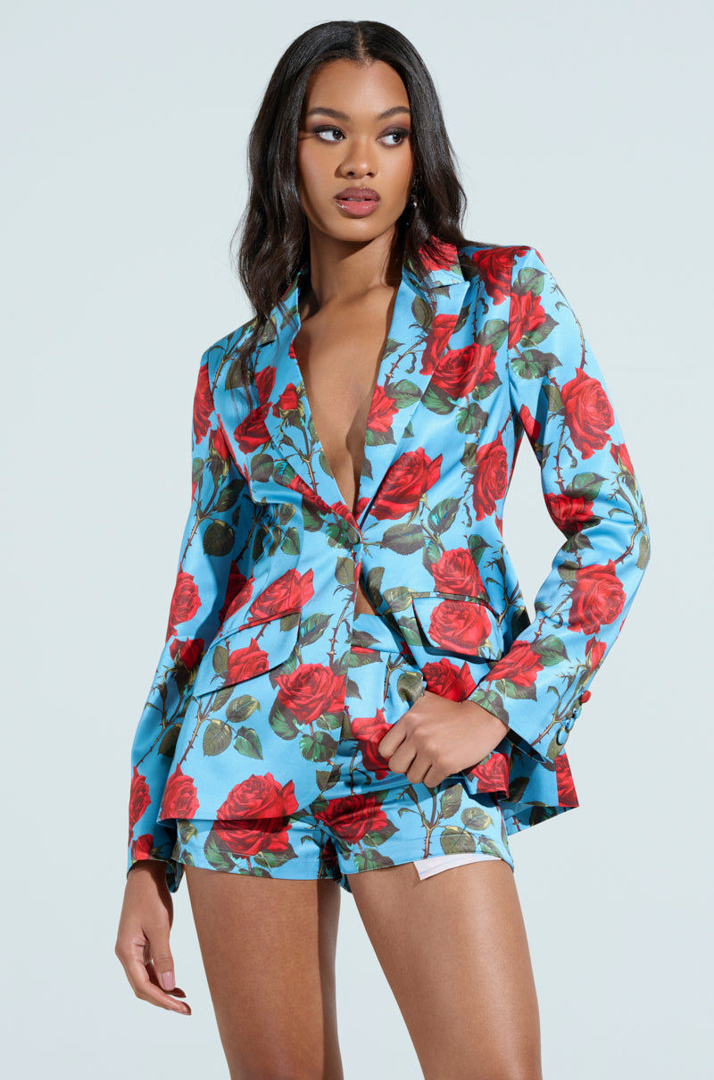 Dressed in the Dozen Roses Satin Blazer, a person exudes charisma with its colorful floral pattern on a blue background. Their long hair enhances the look, matching perfectly with stylish shorts for a vibrant ensemble.