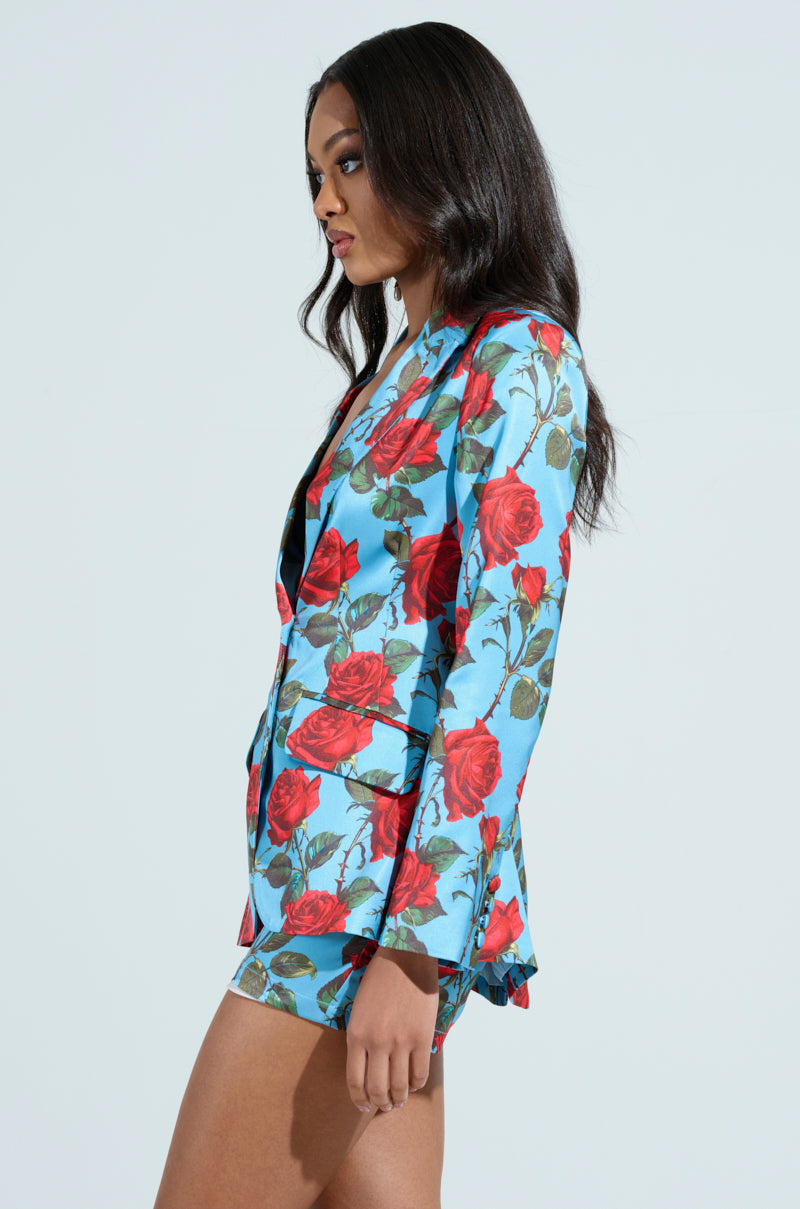 A person with long dark hair wears the DOZEN ROSES SATIN BLAZER, featuring a vibrant blue base adorned with red rose prints. They are shown from the side against a plain light background.