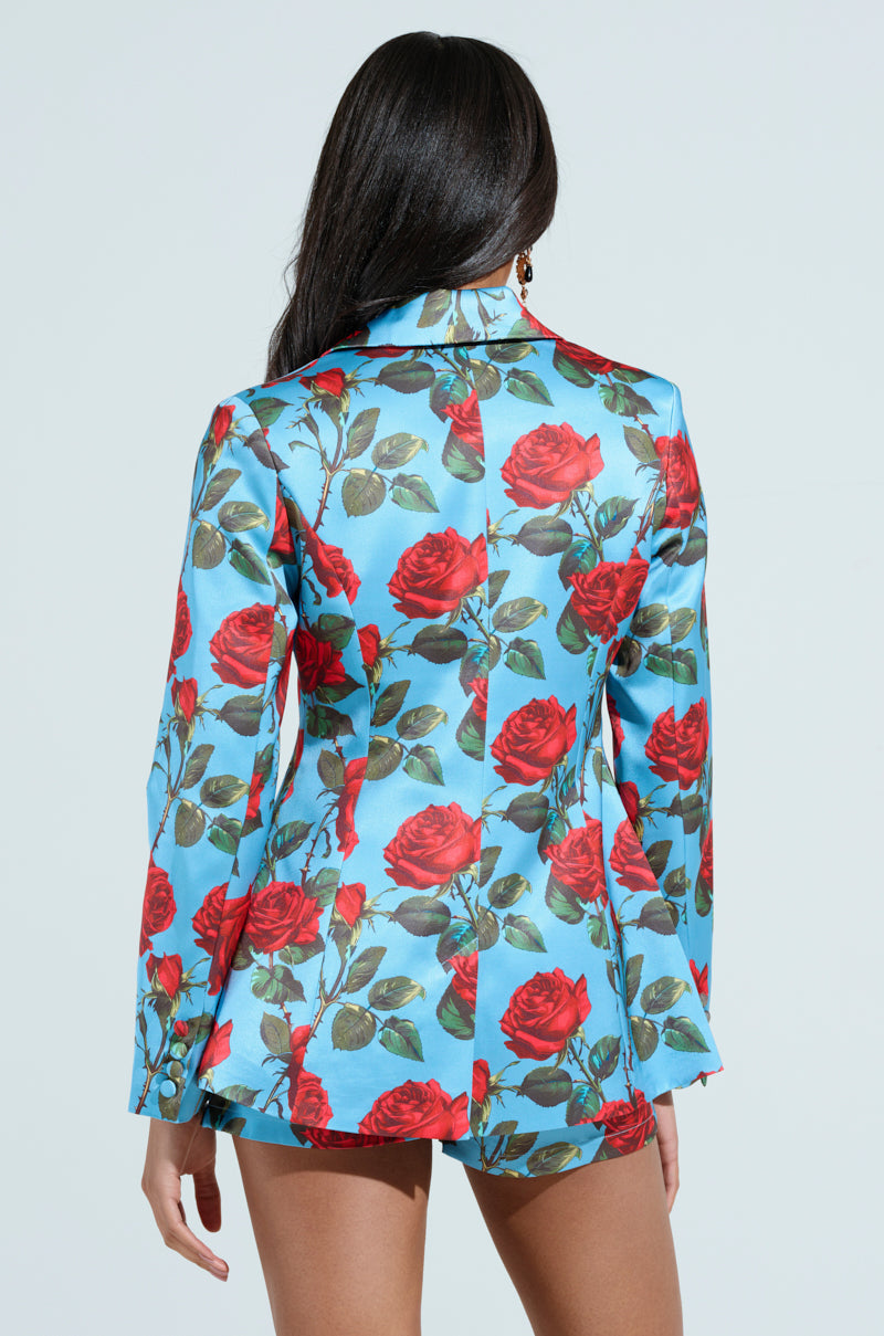 A person with long dark hair, back to the camera, wears the DOZEN ROSES SATIN BLAZER featuring red roses and green leaves. The tailored fit and buttoned cuffs offer an elegant silhouette against a light grey background.