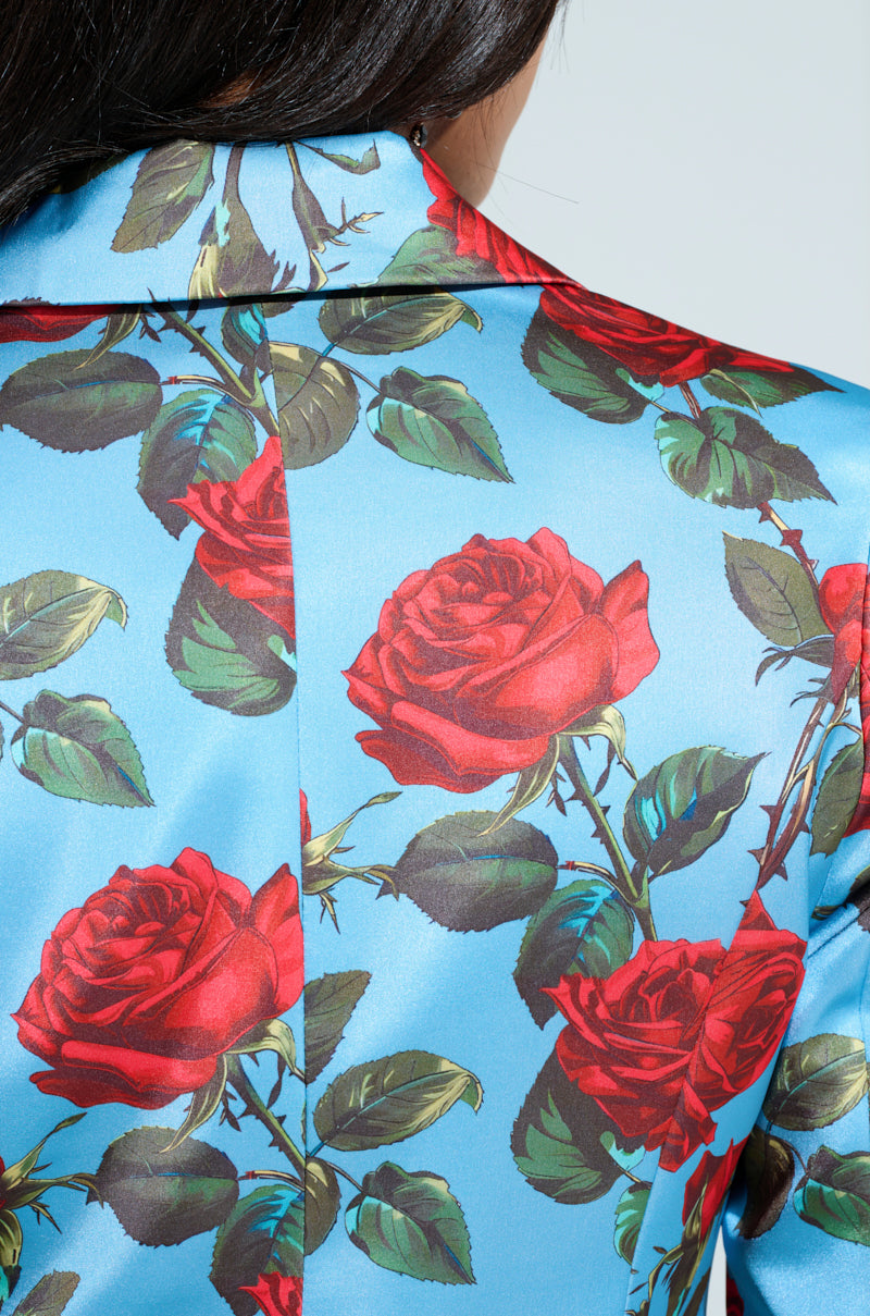 Close-up of a person wearing the DOZEN ROSES SATIN BLAZER, showcasing a vibrant red rose and green leaf pattern on blue fabric. The focus is on the back, highlighting its detailed floral design.
