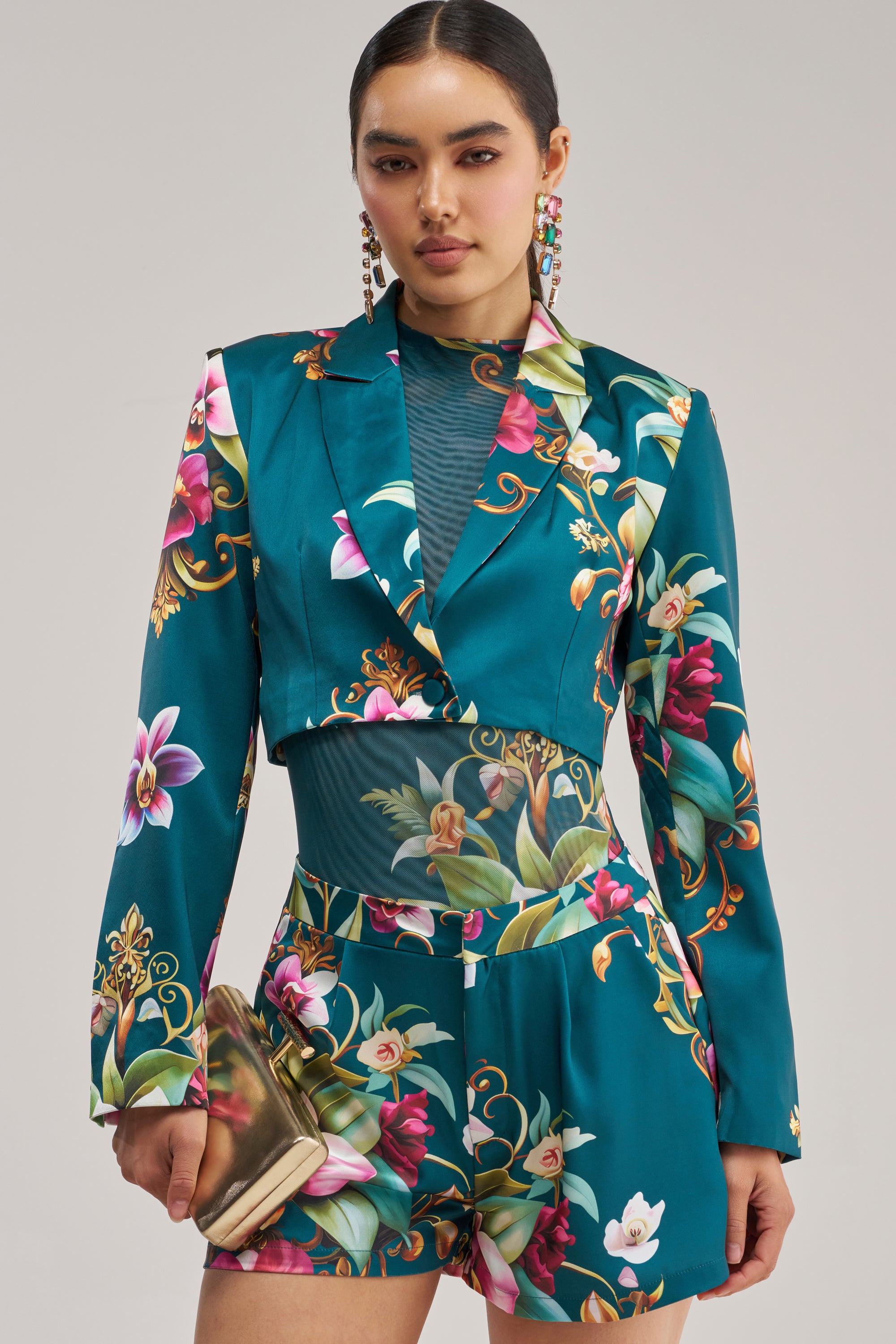 A woman wearing the ISLAND TIME CROPPED SATIN BLAZER in teal floral print with matching shorts and a sheer teal top underneath holds a gold clutch and wears statement earrings, posing effortlessly against a neutral background.