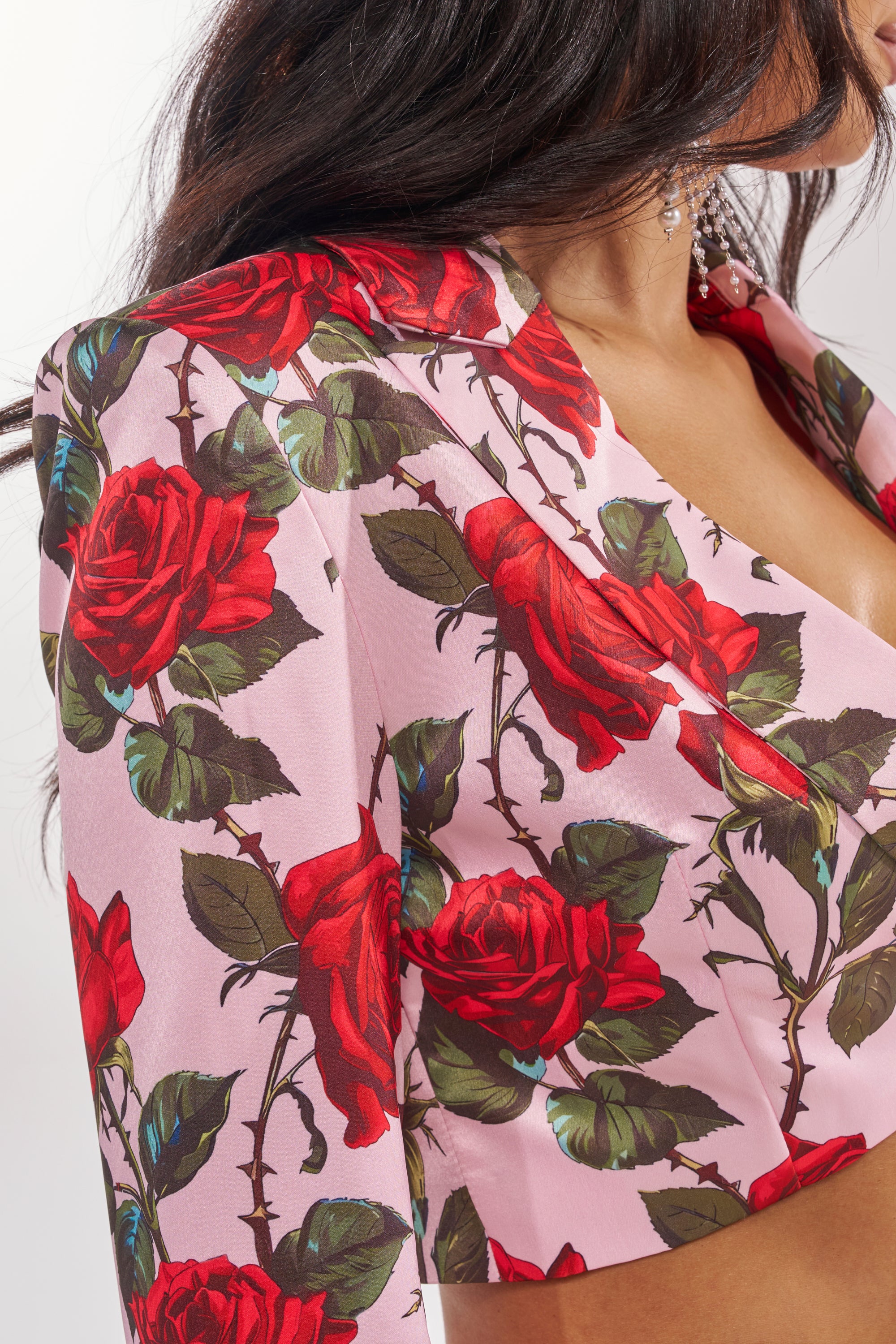 A woman wears the DOZEN ROSES SATIN BLAZER IN LIGHT PINK, cropped to reveal her upper body. Her wavy dark hair frames crystal drop earrings, and the bold rose-print blazer draws attention to her chest. 10