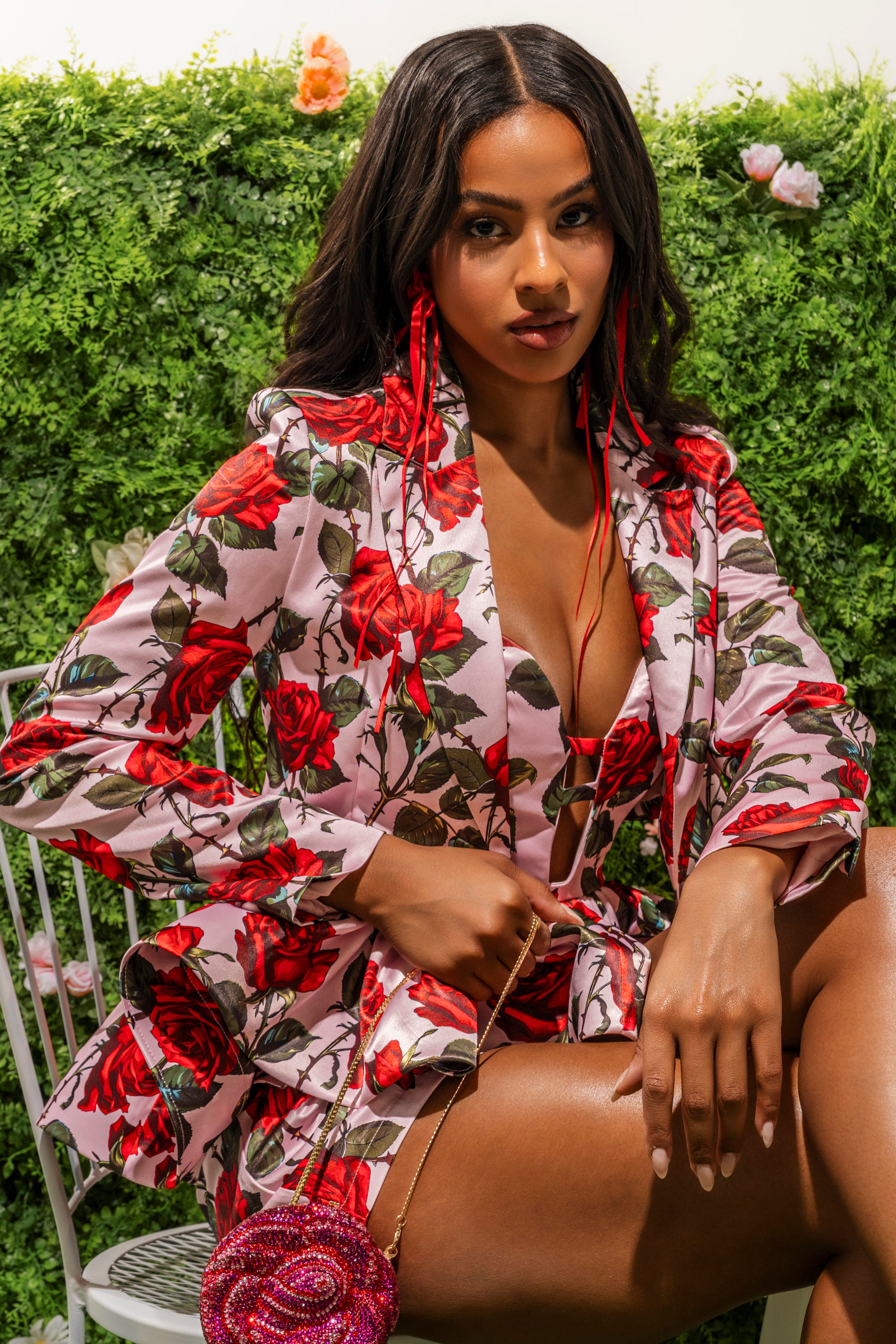 A woman with long dark hair wears the DOZEN ROSES SATIN BLAZER IN LIGHT PINK, styled with matching earrings and a round rose-shaped purse, seated before a lush green backdrop and exuding confidence. 1