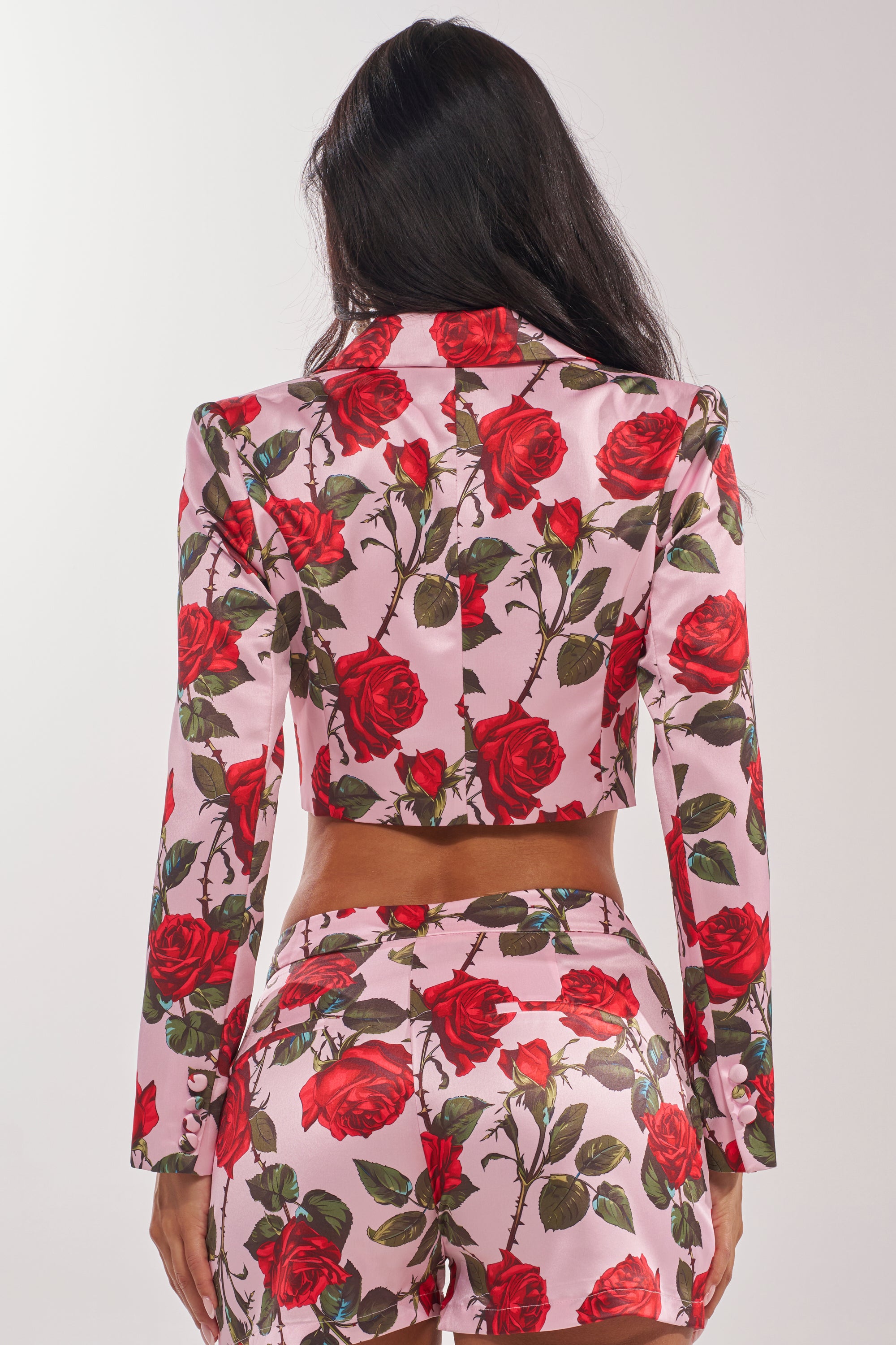 A woman with long dark hair faces away, wearing the DOZEN ROSES SATIN BLAZER IN LIGHT PINK, a rose-print blazer and matching pants adorned with vibrant red roses and green stems, set against a plain white background. 7