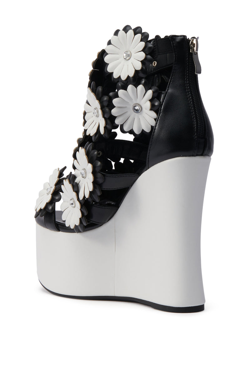 The AZALEA WANG FLOWER EMBELLISHED WEDGE SANDAL IN BLACK features black leather straps with bold white floral appliques and a thick white wedge heel, adding both height and standout style.