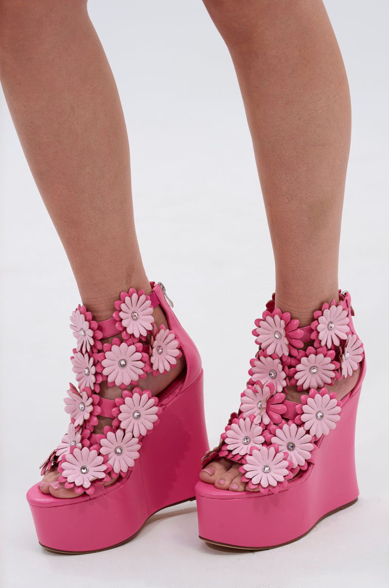 A person wears the AZALEA WANG FLOWER EMBELLISHED WEDGE SANDAL IN PINK, featuring bright faux leather and bold 3D floral appliqués, standing on a light background.